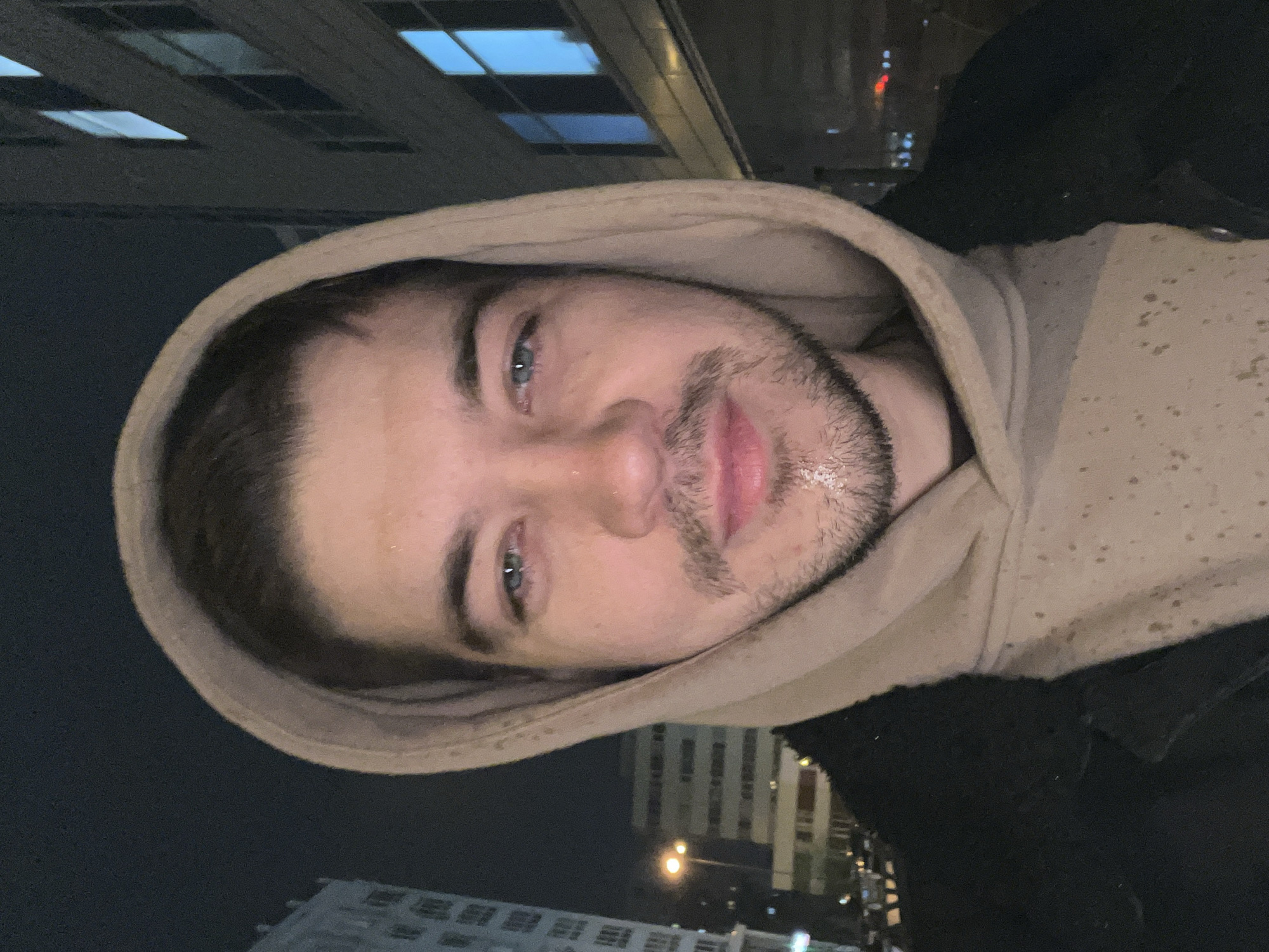 A man with light skin, blue eyes, and a light beard is wearing a beige hoodie and black jacket. It is nighttime. The man is outside in what appears to be Yekaterinburg, Russia, based on the architecture of buildings with visible grid patterns of windows. A street light is visible in the background.