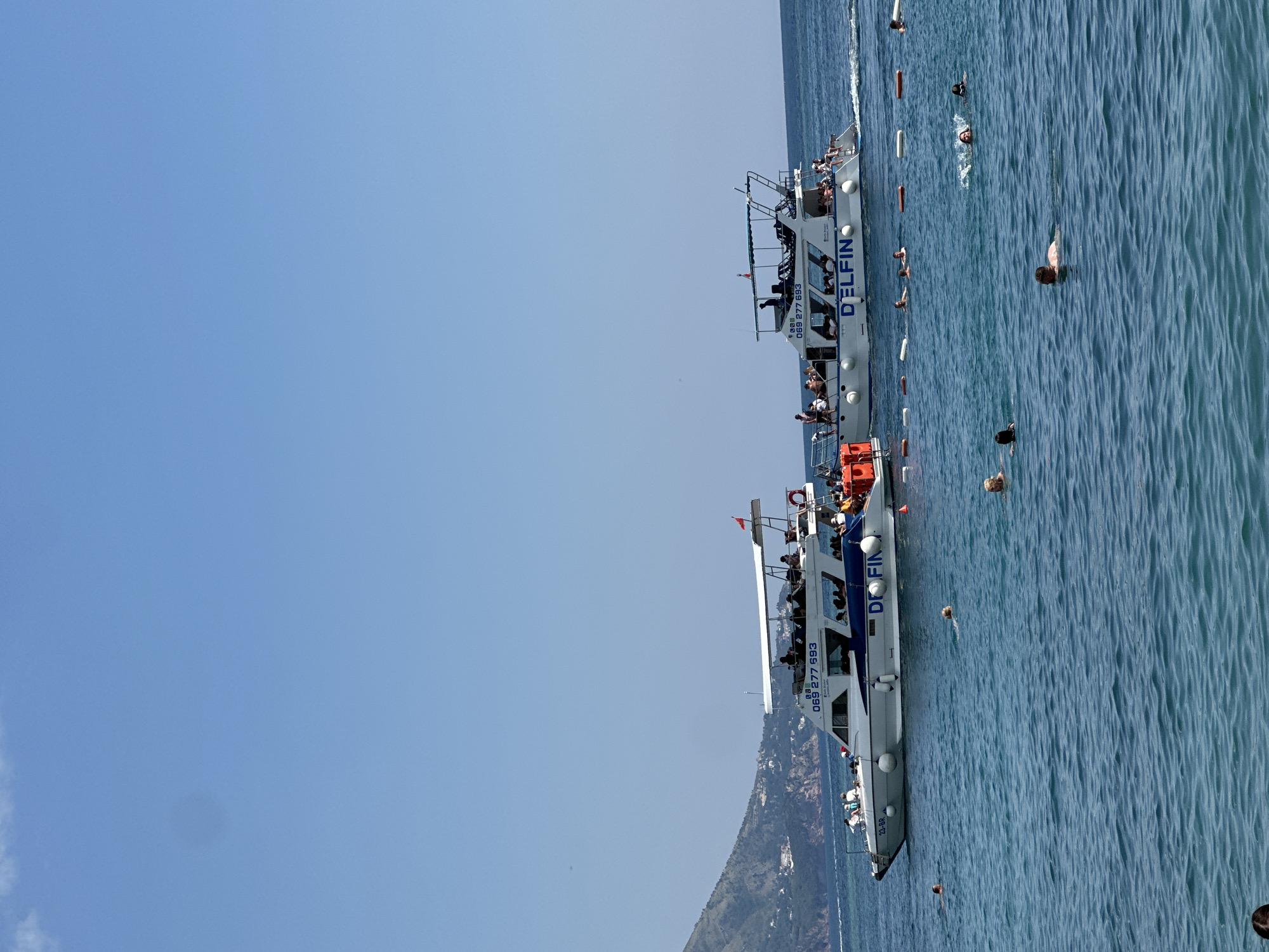 This is a photograph taken from a boat on a bright, clear day. The sky is a vibrant blue with no clouds. The sea is a deep blue with moderate ripples, indicating a calm but not completely still body of water. Two white boats, identified by the text "DELFIN" and "DELFINO" on their sides, are the primary objects in the image. Both boats appear to be pleasure or tour boats, with multiple passengers visible on their decks. The passengers are dressed in casual summer clothing, suggesting a warm climate. Some individuals are standing, while others are seated. The boats have a similar design, featuring multiple levels and railings for safety. One of the boats has the registration number "069 277 693" visible.
The activity taking place is likely a boat tour or a leisure excursion. Several people are in the water around the boats, swimming and enjoying the sea. Their presence suggests a recreational setting, possibly a popular tourist spot. In the background, a lush green, mountainous coastline is visible, with houses and structures built on the slopes. This geographical feature provides a strong cue about the location. The overall atmosphere appears relaxed and enjoyable, with people engaging in water activities under the bright sun. The time of day is clearly daytime due to the strong sunlight and clear sky, and the weather is fair and warm. The presence of the "DELFIN" and "DELFINO" text, coupled with the mountainous coastal scenery, strongly suggests a location in the Adriatic Sea, consistent with Budva, Montenegro.