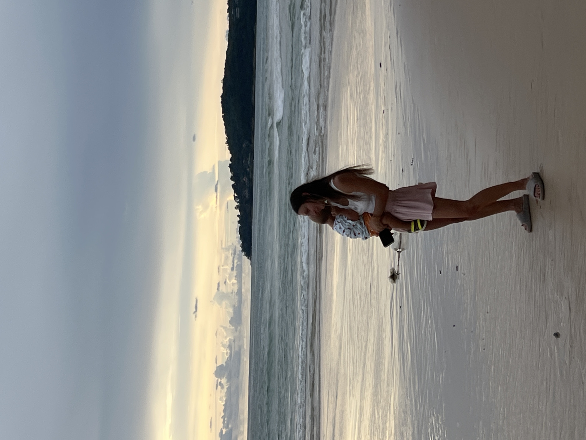 A woman and a baby are standing on a beach in Choeng Thale, Thailand, with the ocean and sky in the background. The woman is wearing a white top and a pink skirt, and she is holding the baby. The baby is wearing a patterned shirt and orange shorts. The beach is sandy, and there are waves in the ocean. The sky is cloudy, with hints of yellow and orange from the setting sun. The overall mood of the image is peaceful and serene.