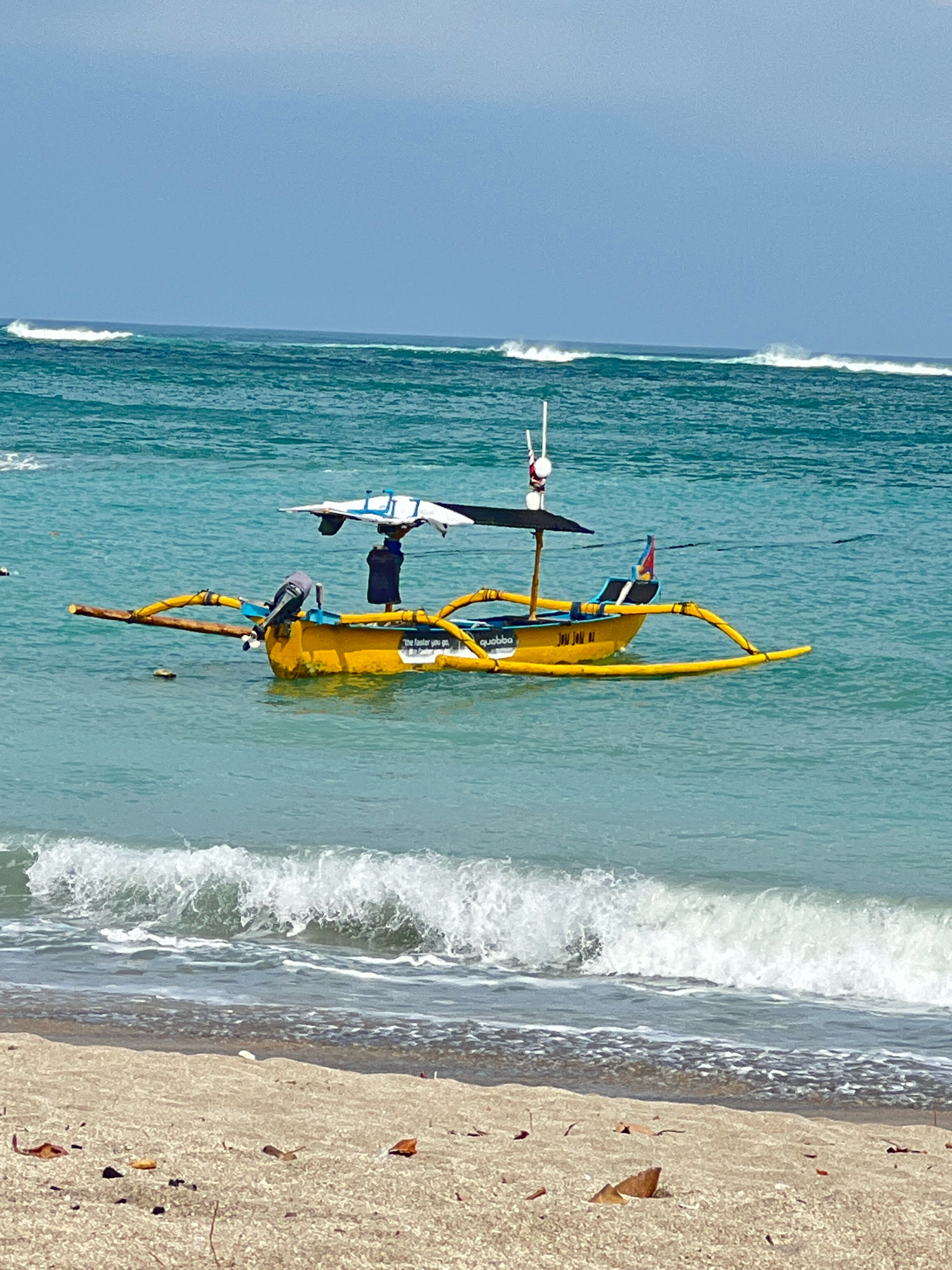 A yellow outrigger boat is moored in shallow turquoise water off the coast of Kuta, Indonesia. The boat has a dark canopy providing shade over a person standing within. An outboard motor is attached to the stern. White-capped waves are breaking on a sandy beach in the foreground, and further out, larger waves roll towards the shore under a hazy blue sky.