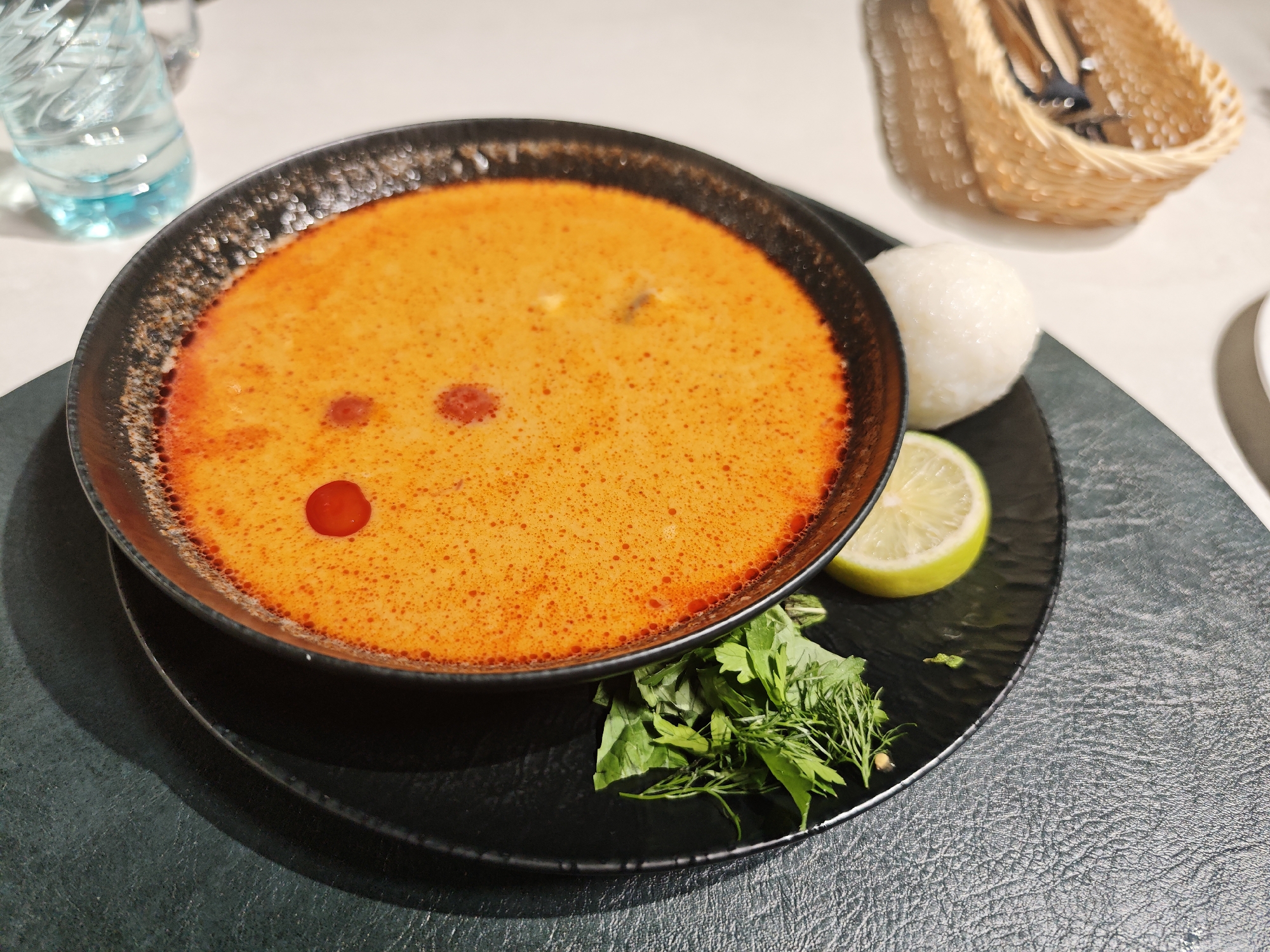 A close-up shot captures a bowl of bright orange soup, likely Tom Yum, served with a side of white rice, a lemon wedge, and fresh green herbs. The soup is presented in a dark, textured bowl, which sits on a black plate. The plate is placed on a textured, dark green placemat. In the background, a glass bottle of water with a blue tint is partially visible, along with a wicker basket holding cutlery. The setting appears to be a restaurant, possibly in Glodeni, Moldova, given the context. The lighting is warm, casting shadows on the placemat. There are no people visible in the frame, and the focus is solely on the meal.