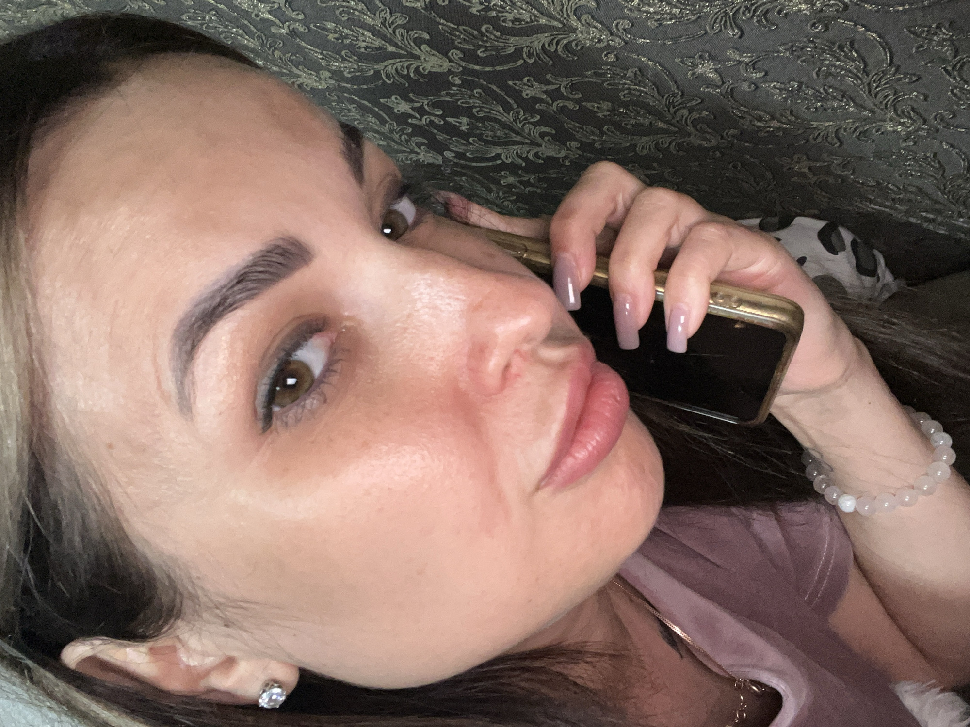 This is a close-up, high-angle selfie of a woman lying down and talking on a gold-rimmed smartphone. The woman has brown eyes, defined eyebrows, and full lips. She is wearing a dusty rose colored shirt and a diamond stud earring in her left ear. She also has a beaded bracelet on her right wrist. The background appears to be a textured, dark fabric, possibly a couch or a blanket. The lighting suggests it is daytime, but the setting is indeterminate. The woman's expression is somewhat neutral, with her lips slightly pursed as if in conversation. The focus is on her face and the phone.