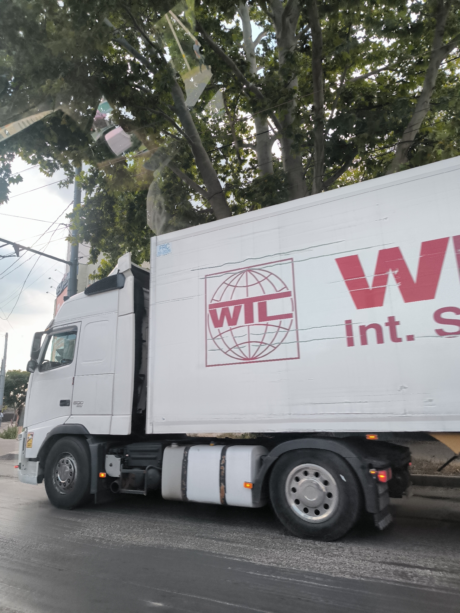 A white Volvo semi-truck with a white trailer is driving on a city street in Chișinău, Moldova. The trailer has a red logo with "WIL" in the center of a globe, and the text "Int. S" below it. The truck has the number "500" on its side. Trees with green leaves are visible above the truck. The sky is cloudy. The road surface is dark and appears wet or dirty. There are power lines and some buildings in the background. A person is visible walking on the sidewalk in the distance on the left side of the image.