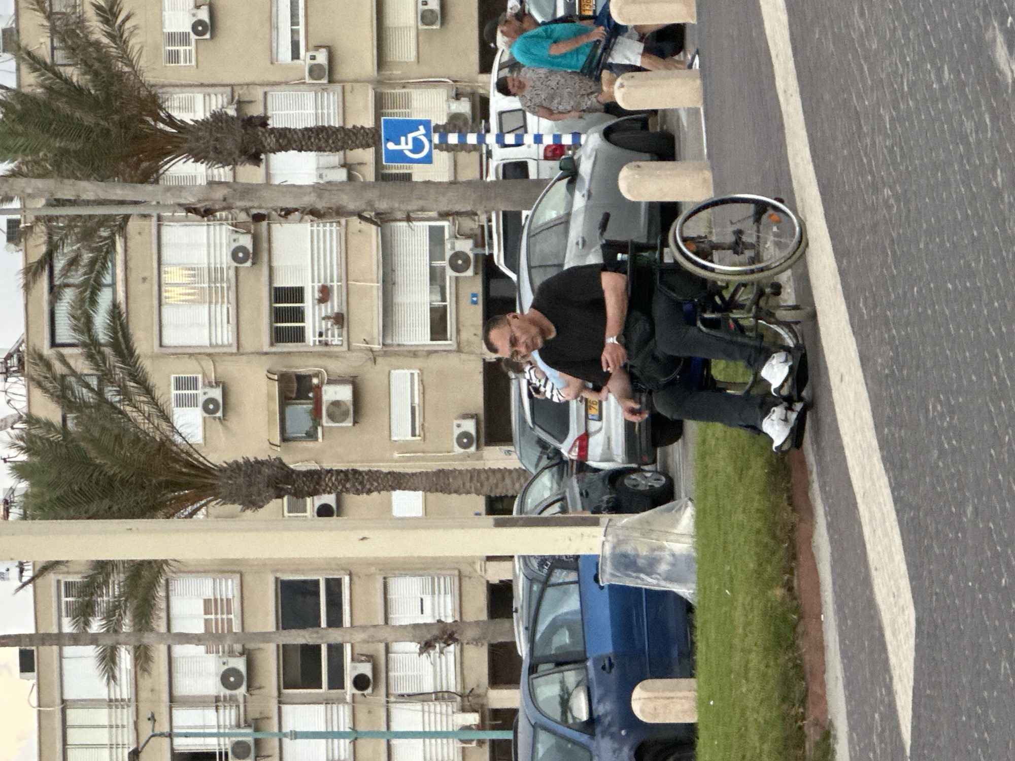 The image depicts a street scene in Haifa, Israel, with a focus on a man in a wheelchair and the surrounding environment. The man is wearing a black t-shirt and dark pants, and he appears to be pushing himself forward in the wheelchair. He is looking towards the viewer and seems to have a neutral or slightly concerned expression.
Behind the man, a building with several windows and air conditioning units is visible. The building's facade is a light beige color. A palm tree with lush fronds extends from the left side of the image, partially obscuring the building. In the foreground, a paved road with a white line marking is present, suggesting a public thoroughfare. A grassy area borders the road, and a curb is visible.
In the background, several vehicles are parked. A gray car is partially visible to the right of the man in the wheelchair, and a blue vehicle is further in the background. Other people are also present in the scene, though they are less distinct. One person wearing a blue t-shirt and shorts is sitting in a wheelchair, and another individual in a patterned shirt is standing. A blue sign with a white wheelchair symbol is mounted on a pole, indicating a disabled parking spot or accessible area.
The lighting suggests it is daytime, possibly late afternoon or early evening, given the shadows cast on the road. The weather appears to be clear. The overall impression is a snapshot of everyday life on a street in an urban environment, with an emphasis on accessibility.