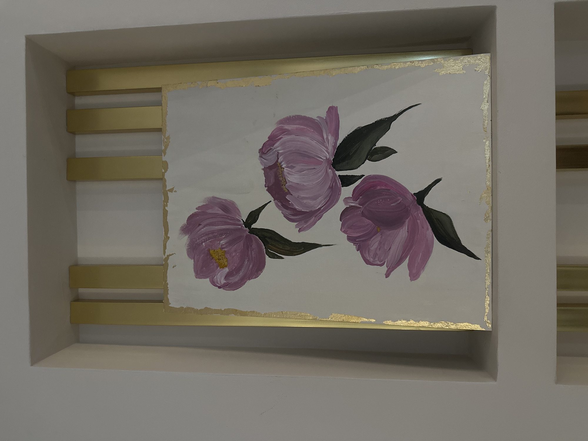 An artwork depicting three purple or pink flowers with dark green leaves on a light background is displayed within a recessed niche in a wall. The painting itself features a rough, textured gold border, suggesting gold leaf or paint applied to its edges. The flowers, possibly peonies, are rendered in a loose, painterly style. The central flower is slightly larger and more open than the two smaller ones.
The painting is supported by a series of horizontal, gold-colored metal bars or shelves. Three such bars are visible to the left of the painting, extending from the left wall of the niche to partially beneath the artwork. Similar bars are partially visible to the right, suggesting a continuous support system. The walls of the niche and the surrounding room are a plain, light neutral color, likely off-white or light gray. The overall presentation suggests an intentional and decorative display of the artwork in an indoor setting. There are no people visible, nor any discernable text within the image. The lighting appears to be artificial, casting soft shadows within the recessed area.