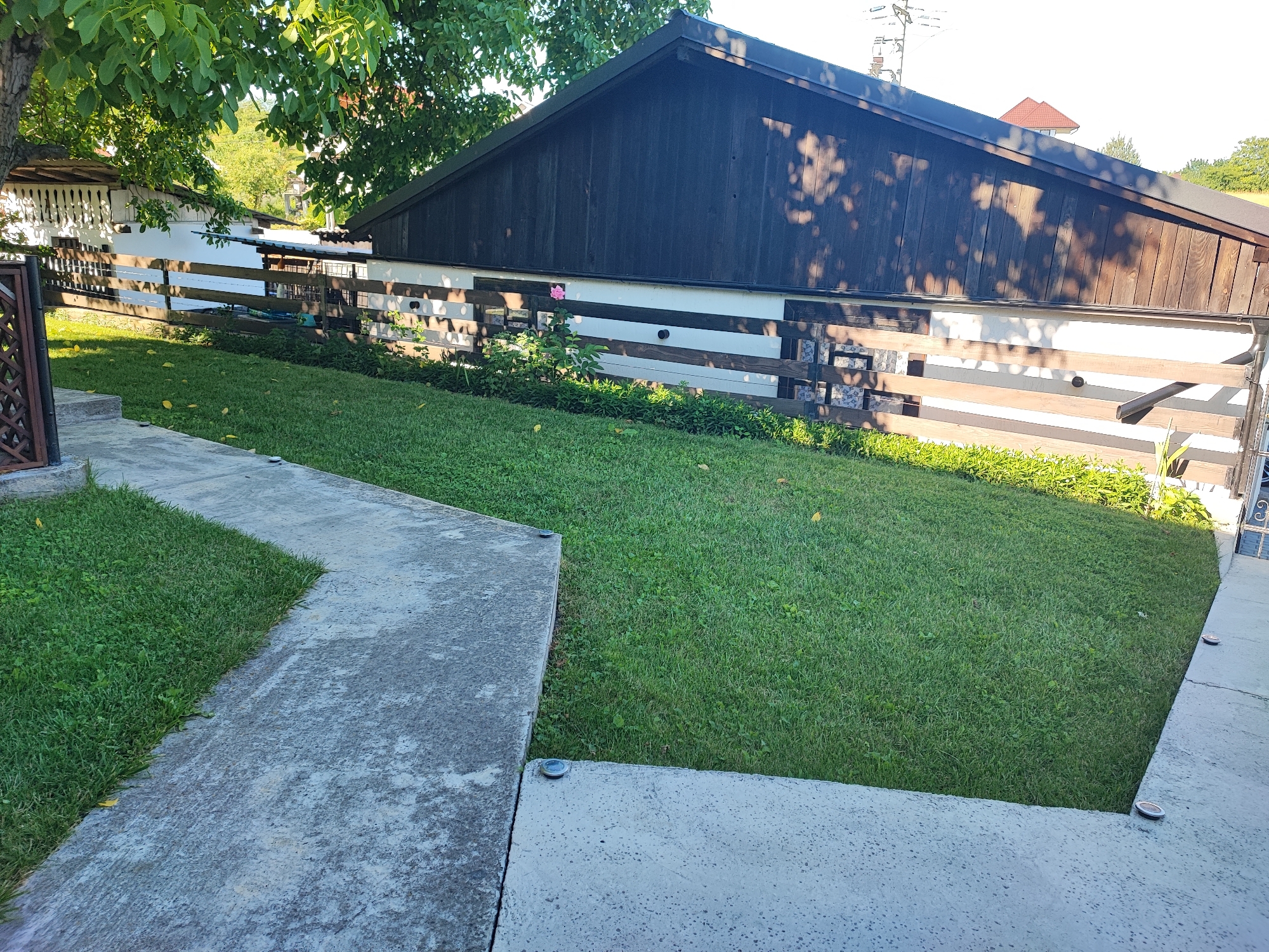 The image depicts a backyard scene in Târgu Neamț, Romania. In the foreground, a concrete path curves to the left, bordering a well-maintained green lawn that slopes upwards. Beyond the lawn, a wooden fence separates the yard from a building. The building features a dark wooden roof and a light-colored wall with horizontal wooden planks. A single pink rose is visible growing near the fence. In the background, trees and a portion of another building with a red roof are visible against a clear sky. The overall impression is that of a peaceful residential setting.