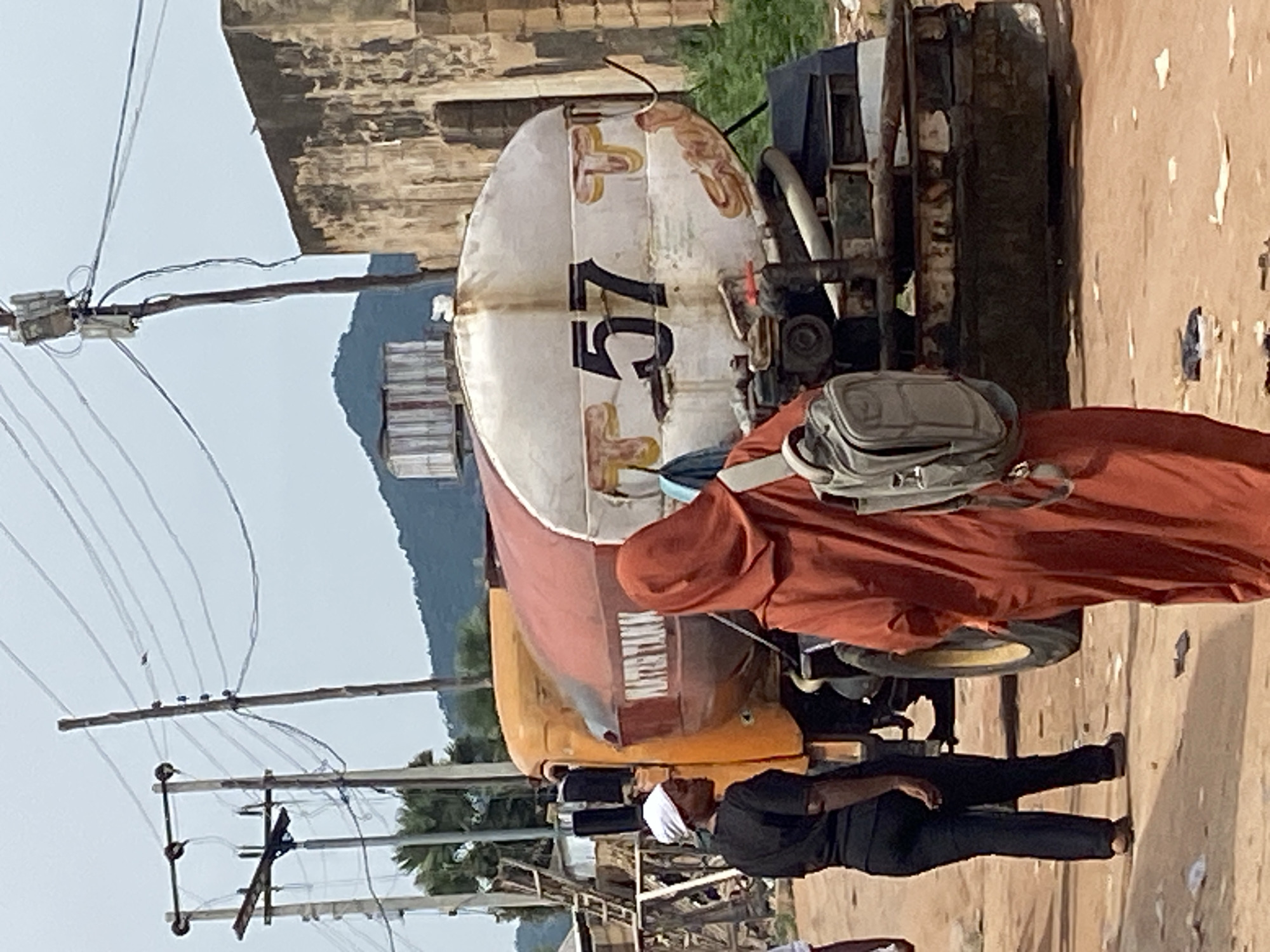 This image depicts a street scene in Kuson, Nigeria, likely during daylight hours. The foreground is dominated by a person wearing a rust-colored hooded garment, with a gray backpack slung over their shoulder. To their left, another person is visible, dressed in black attire and a white head covering, looking upwards.
Behind the individuals is a large, yellow truck with a white cylindrical tank mounted on it. The tank bears the number "57" prominently in black lettering, along with some faded decorative elements. The truck appears to be an older model, with visible signs of wear and tear.
In the background, there are indications of a built environment with structures made of brick. Overhead power lines and utility poles are a significant feature of the scene, suggesting an urban or semi-urban setting. Beyond the immediate structures, a hill or mountain range is visible, and lush green vegetation can be seen in the mid-ground, hinting at a somewhat rural or natural landscape surrounding the populated area. The ground is a dusty, unpaved surface.
The overall impression is one of everyday life in a Nigerian community, with the truck possibly involved in a delivery or service, and the people going about their daily activities. There are no strong cues for time of day beyond it being light, and the weather appears to be clear, though the sky is overcast. The focus on the individuals and the vehicle suggests a moment captured in time rather than a specific event. No legible text other than the "57" on the tank is clearly discernible.