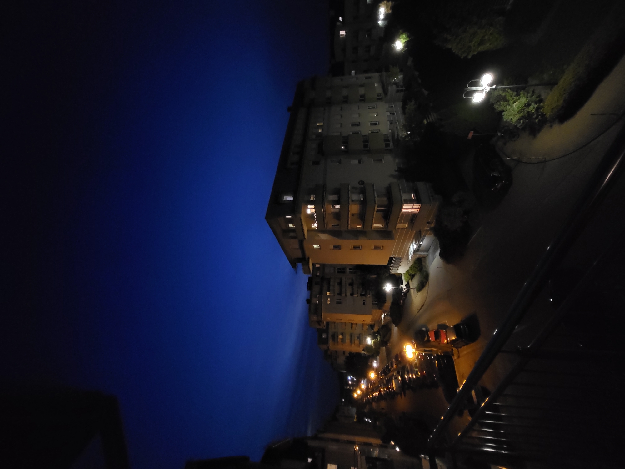 An elevated view looks out over a street at night in an urban area, likely in Europe. The sky is a deep, clear blue, transitioning to black at the top of the frame. Several multi-story apartment buildings line the street, their windows illuminated with warm yellow light, indicating occupancy. A row of parked cars is visible along the side of the street, with the headlights of some cars and streetlights casting bright pools of light. A wrought-iron balcony railing is in the foreground, partially obscuring the view. The scene is peaceful and quiet, with no people visible.