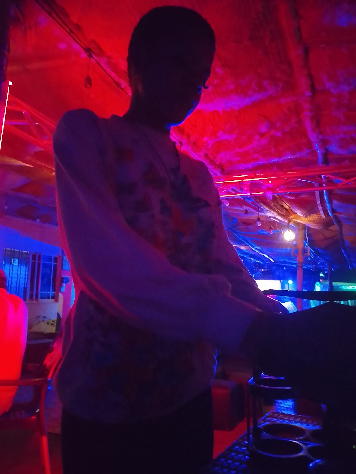 A person is depicted in a vibrant, dimly lit indoor or semi-enclosed space, possibly a club or event venue. The scene is dominated by strong artificial lighting, primarily red from above and blue from various sources, casting the main subject largely in silhouette.
The central subject is a person facing slightly to the right, their head bowed, with their face obscured by shadow. They are wearing a light-colored, long-sleeved top with a subtle pattern visible on the fabric where the light catches it, and dark bottoms. Their hands appear to be resting on or interacting with an object in the right foreground.
In the immediate foreground to the right, a dark, multi-tiered stand or rack is visible, featuring several circular openings, suggesting it holds drinks or other items. Below this, there's a hint of a dark, textured surface, possibly a keyboard or control panel.
The background reveals structural elements, including beams and possibly draped fabric or sheeting, illuminated by the intense red light. Several small, hanging light bulbs are visible amongst these structures. To the far left, a window with vertical bars can be seen on a light-colored wall, behind which an indistinct red object, possibly a chair or part of a furniture piece, stands. The overall setting suggests a lively, nighttime environment with a distinct, colorful ambiance. No visible text is present in the image.