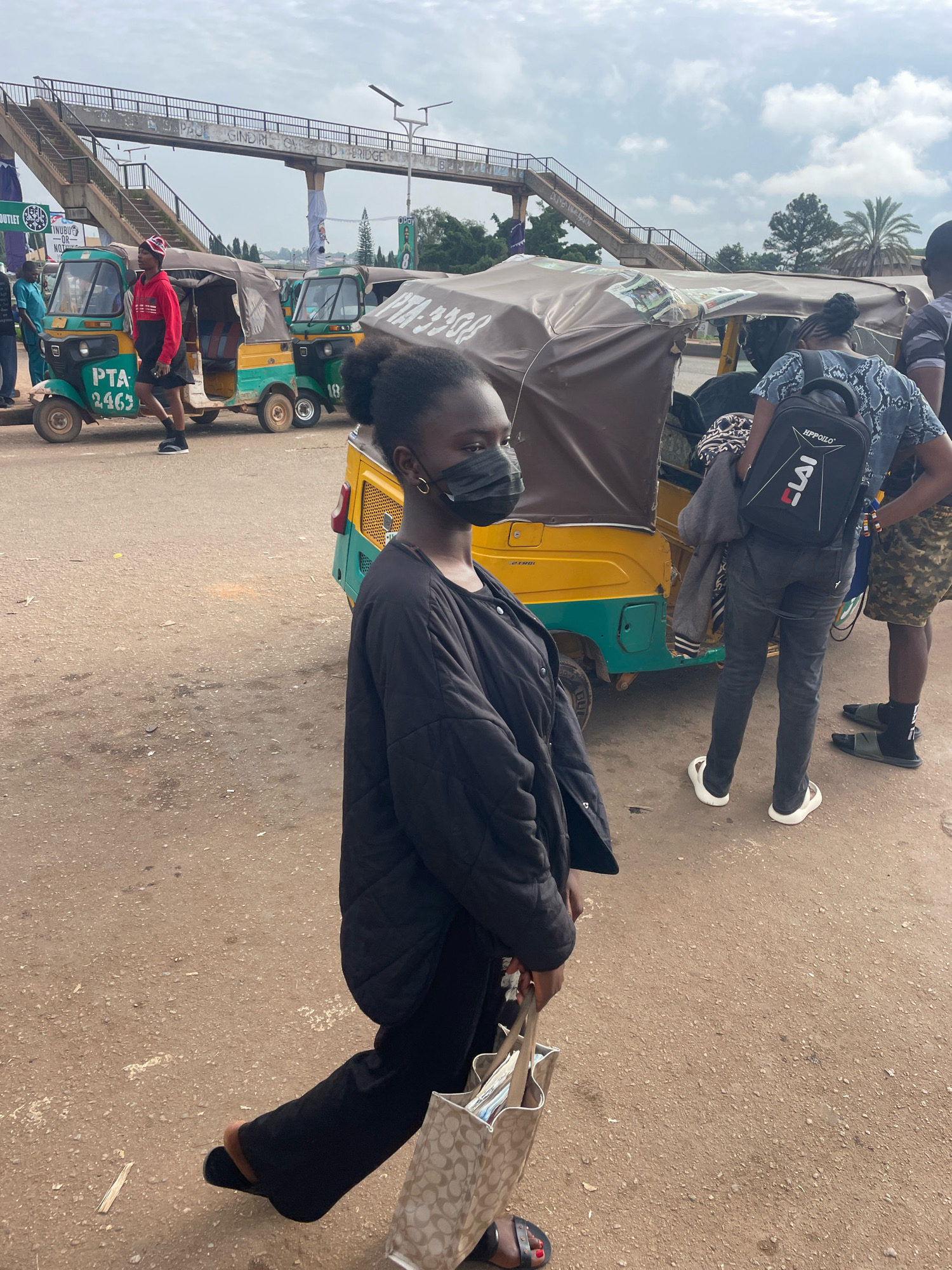The image captures a street scene in Gura Top, Nigeria, under an overcast sky. In the foreground, a young Black woman wearing a black puffer jacket and black pants, with a black face mask, walks from left to right. She carries a beige tote bag with a repeating pattern. Behind her, several auto-rickshaws, also known as "keke napep," are parked or in motion. One auto-rickshaw is predominantly yellow and green, with the number "2463" visible on its side. Another auto-rickshaw, also yellow and green, is partially visible to the right. Further in the background, a pedestrian bridge crosses the street. Several people are present, including a man in a red hoodie and black skirt with a red and white patterned hat, standing next to an auto-rickshaw. Another individual, wearing a black and white patterned shirt and dark pants, carries a black backpack with the word "HIPPOILO" and "LAI" visible. The ground is paved with dirt and gravel. The overall atmosphere suggests a bustling urban environment.