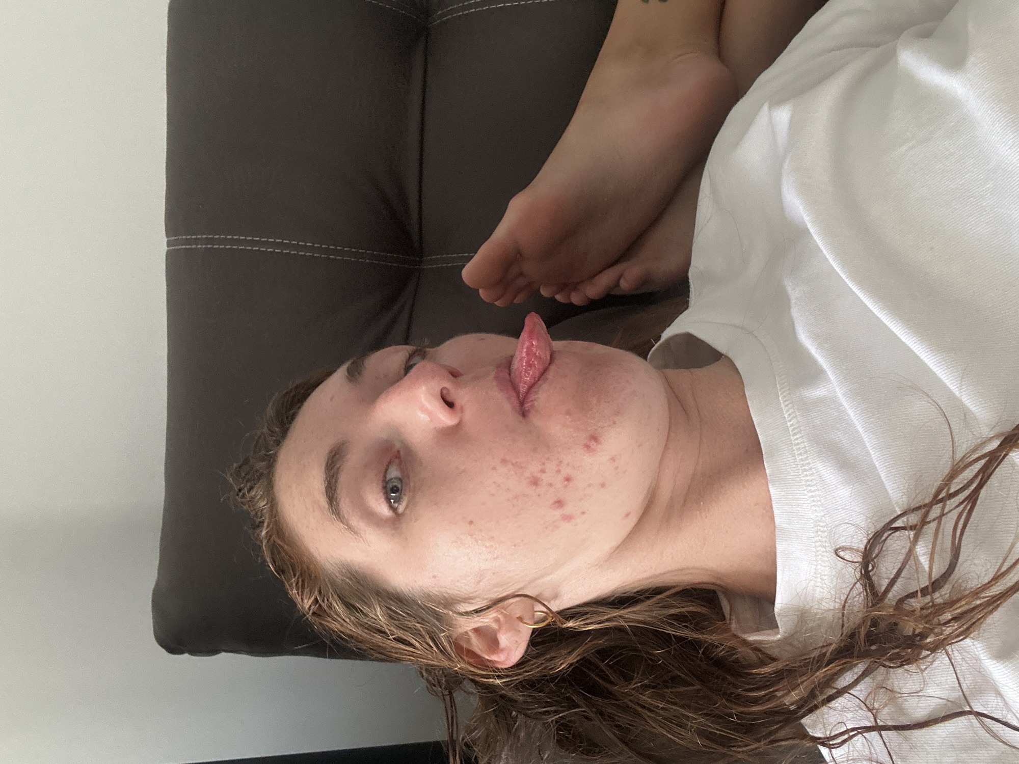 A woman lies on a dark gray cushion, sticking out her tongue. Her feet are resting above her head. She wears a white t-shirt. The location is not discernible and cannot be identified as Baan Chaweng, Thailand, based on the provided image.