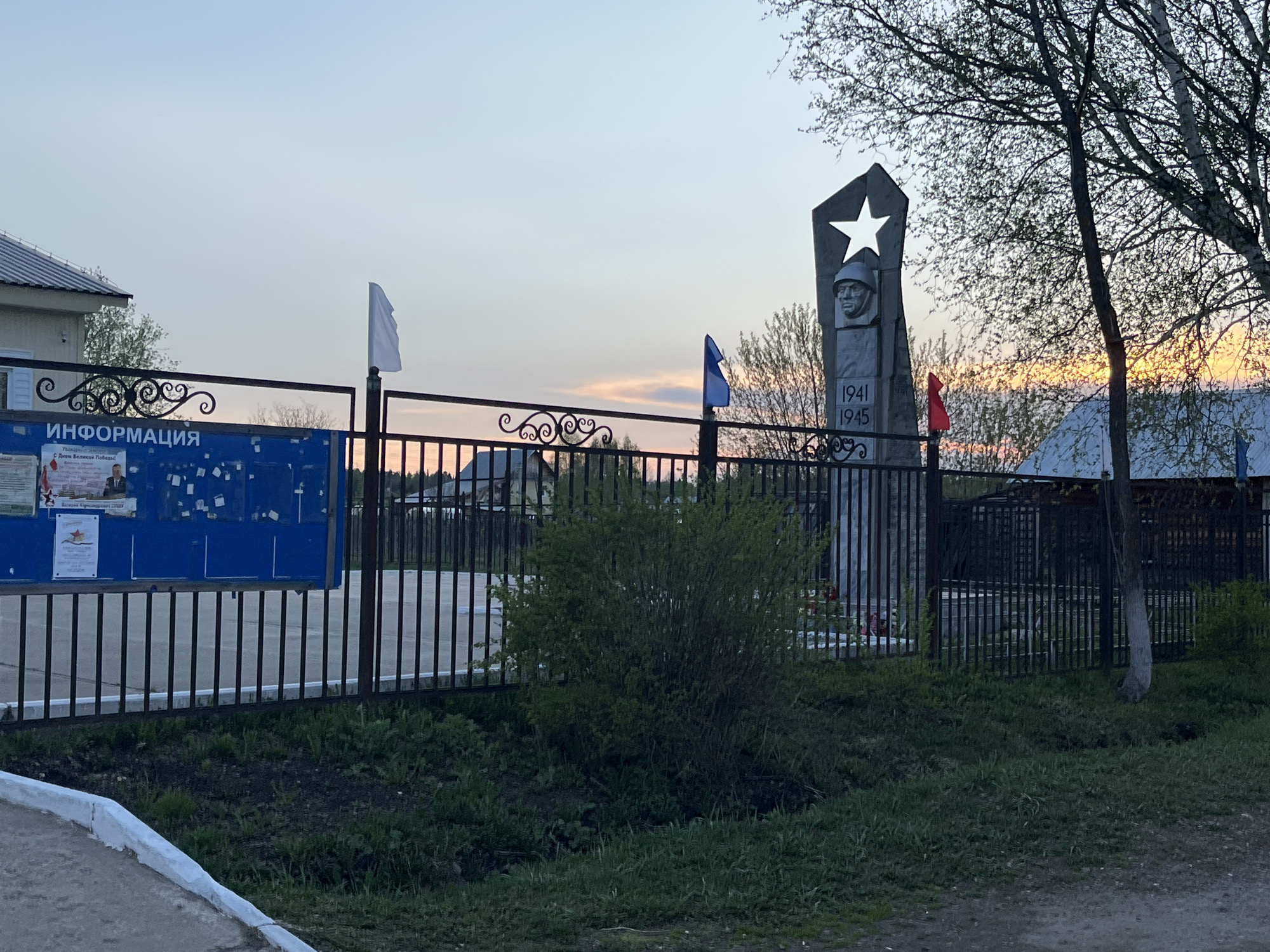 The image shows a memorial in Сосново, Russia, commemorating World War II (1941-1945). The memorial features a gray stone monument with a relief of a soldier's head under a five-pointed star. The monument is surrounded by a black metal fence with Russian flags (white, blue, and red) attached. A blue information board is on the left of the memorial. The area around the memorial is grassy. Buildings and trees are visible in the background under a dusk sky.