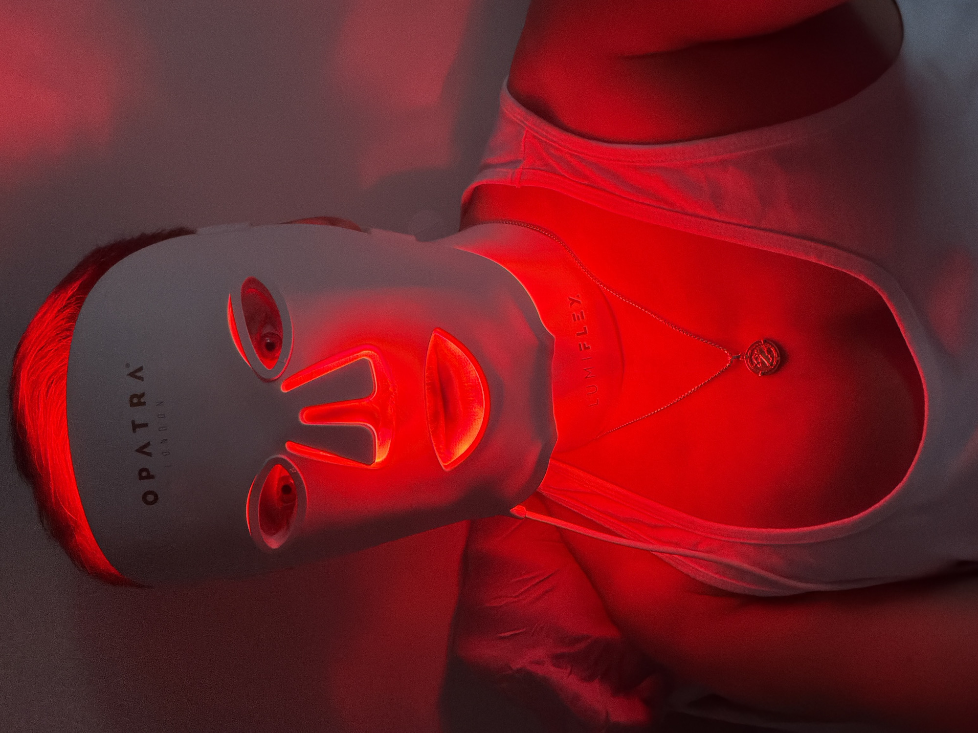 A woman wearing a white tank top lies supine, illuminated by red light emanating from an OPATRA LONDON light therapy mask. She has a gold necklace with a round pendant. The location is not discernible.