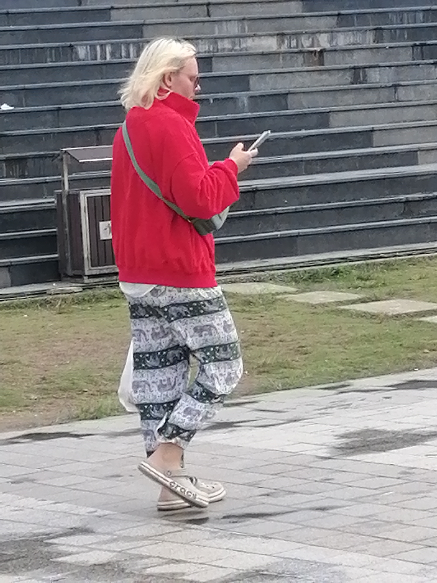 A person with blonde hair and glasses is walking on a paved surface with a stone staircase in the background. They are wearing a red jacket, patterned pants, and light-colored slip-on shoes. A green strap crosses their chest, and they are holding a silver object, possibly a phone, in their right hand. The environment appears to be outdoors in Da Lat, Vietnam, with grass visible in the midground. The lighting suggests it is daytime.