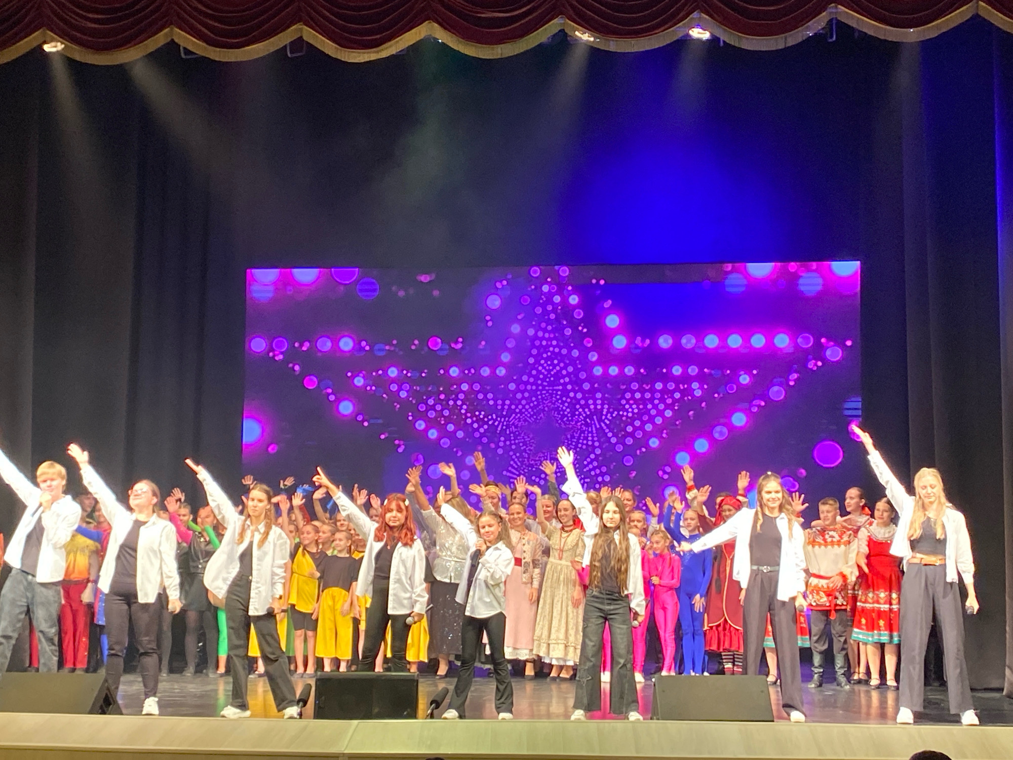 A large group of children and young adults, dressed in a variety of performance costumes including traditional Russian attire, are gathered on a brightly lit stage in front of a large LED screen displaying abstract purple and pink lights and patterns. The performers are a mix of males and females, some holding microphones, and many are waving their hands. The stage is framed by dark curtains and a decorative red and gold valance at the top. The overall atmosphere suggests a performance or concert is taking place. The specific location within Ivanteyevka, Russia is not discernible from the image alone.