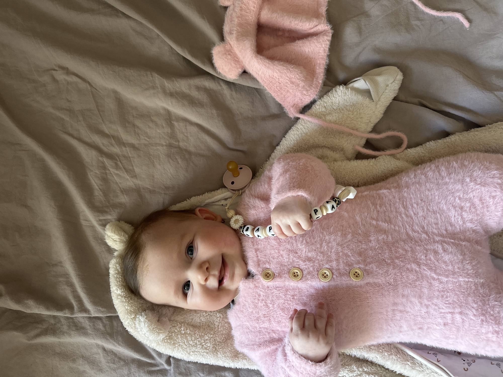 A baby wearing a pink fuzzy onesie with wooden buttons and a white fluffy hood with ears is lying on a light brown sheet. A pink fuzzy hat and a pacifier are next to the baby. The scene is likely indoors, but the city and location of Chaikovsky, Russia, are not evident from the image.