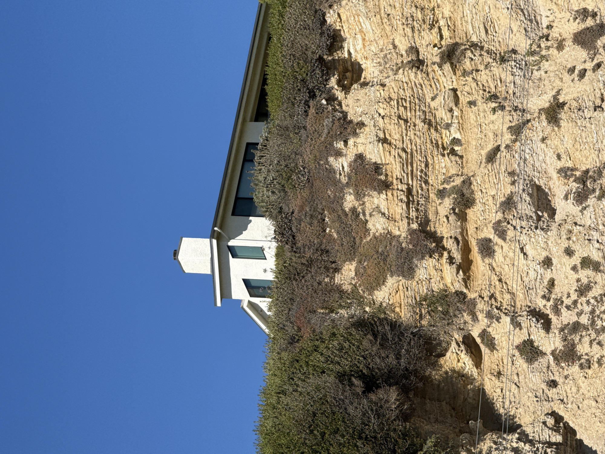 The image shows a white house with a dark roof, a chimney, and large windows, situated on a cliff overlooking the ocean. The cliff is covered in green brush and dry vegetation, with layers of rock visible. The sky is a clear blue. The scene is likely located in Malibu, California, a coastal city known for its dramatic cliffs and ocean views. The house appears to be a modern design, built to take advantage of the scenic surroundings. There are no people or any other living creatures visible in the image. The overall impression is one of tranquility and natural beauty, with the man-made structure harmoniously integrated into the landscape.