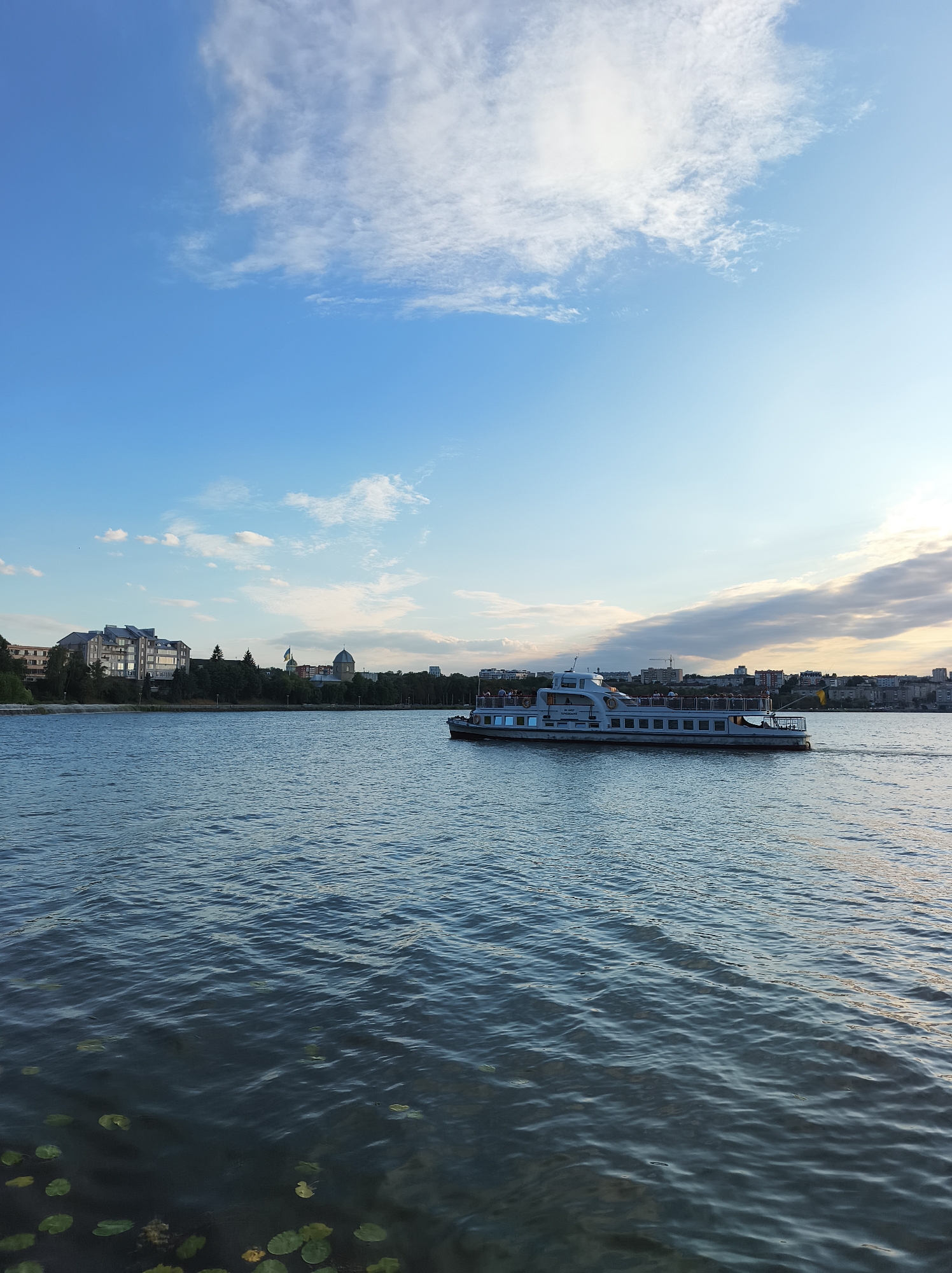 The image captures a serene scene on the Ternopil Lake in Ternopil, Ukraine. A white passenger boat, carrying several people, cruises across the calm, blue water. The boat is positioned in the right half of the frame. The left side of the image features a cityscape with modern buildings and trees lining the shore, with a domed building and a flag visible in the distance. The sky above is a vibrant blue, with scattered white clouds, some tinged with the warm hues of the setting sun. Lily pads are visible floating on the water in the lower left corner of the image.