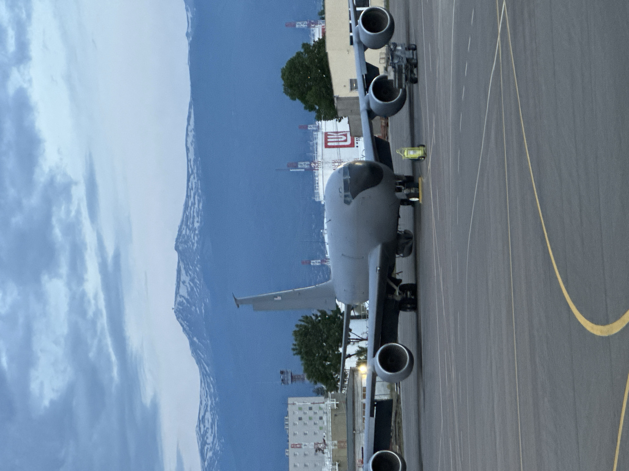 A gray military aircraft sits on an airport tarmac. The aircraft appears to be a KC-135 Stratotanker. Mountains are visible in the background. Buildings, including one with a red logo, are also visible. The tarmac has yellow guide lines. A small yellow vehicle sits near the aircraft.
It is not possible to determine the location as the city of Sofia, Bulgaria.