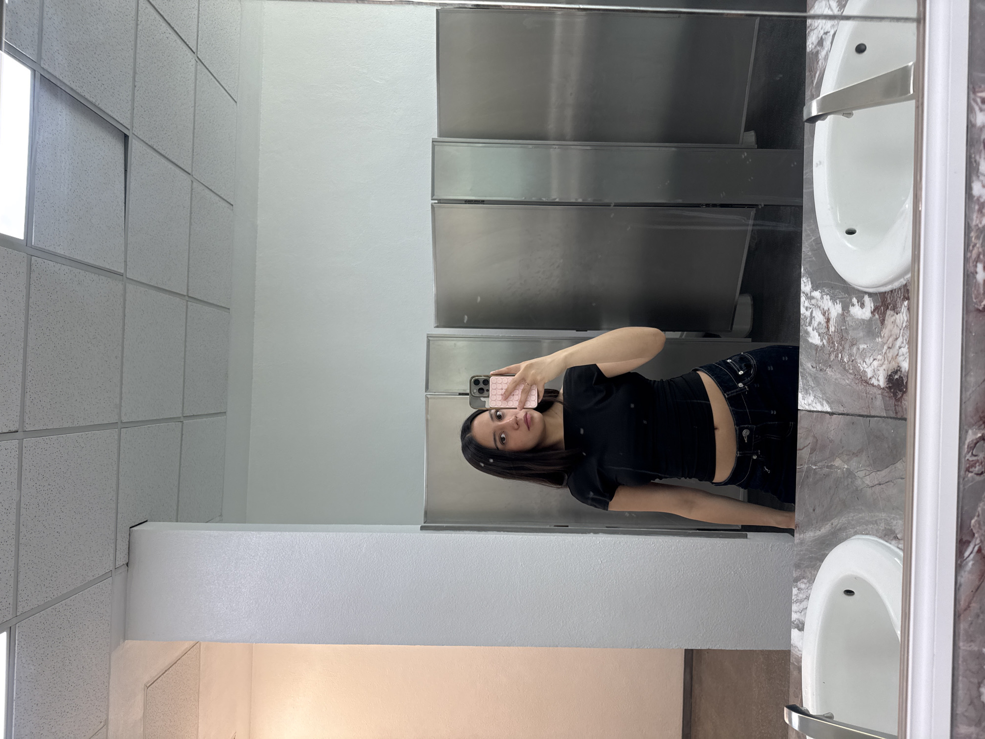 A woman in a black shirt and blue jeans stands in a public restroom, taking a selfie in the mirror with her pink-cased smartphone. The restroom features tiled ceilings, white walls, stainless steel stalls, and marbled countertops with white porcelain sinks. The location is likely within the city of Puebla, Mexico.