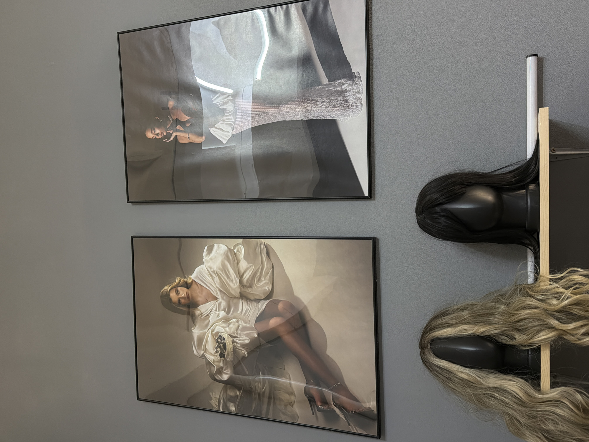 The image shows a gray wall with two framed photographs hanging on the left. The top photograph depicts a person in a silver and gray outfit lying down. The bottom photograph depicts a person in a white outfit reclining on a surface. To the right of the photographs are two black mannequin heads mounted on a wooden and white structure. One head has long, dark hair, and the other has long, blonde hair. The environment suggests an interior space, likely a studio or store. The location is not evident from the image and cannot be determined as Kazan, Russia, with the information given.