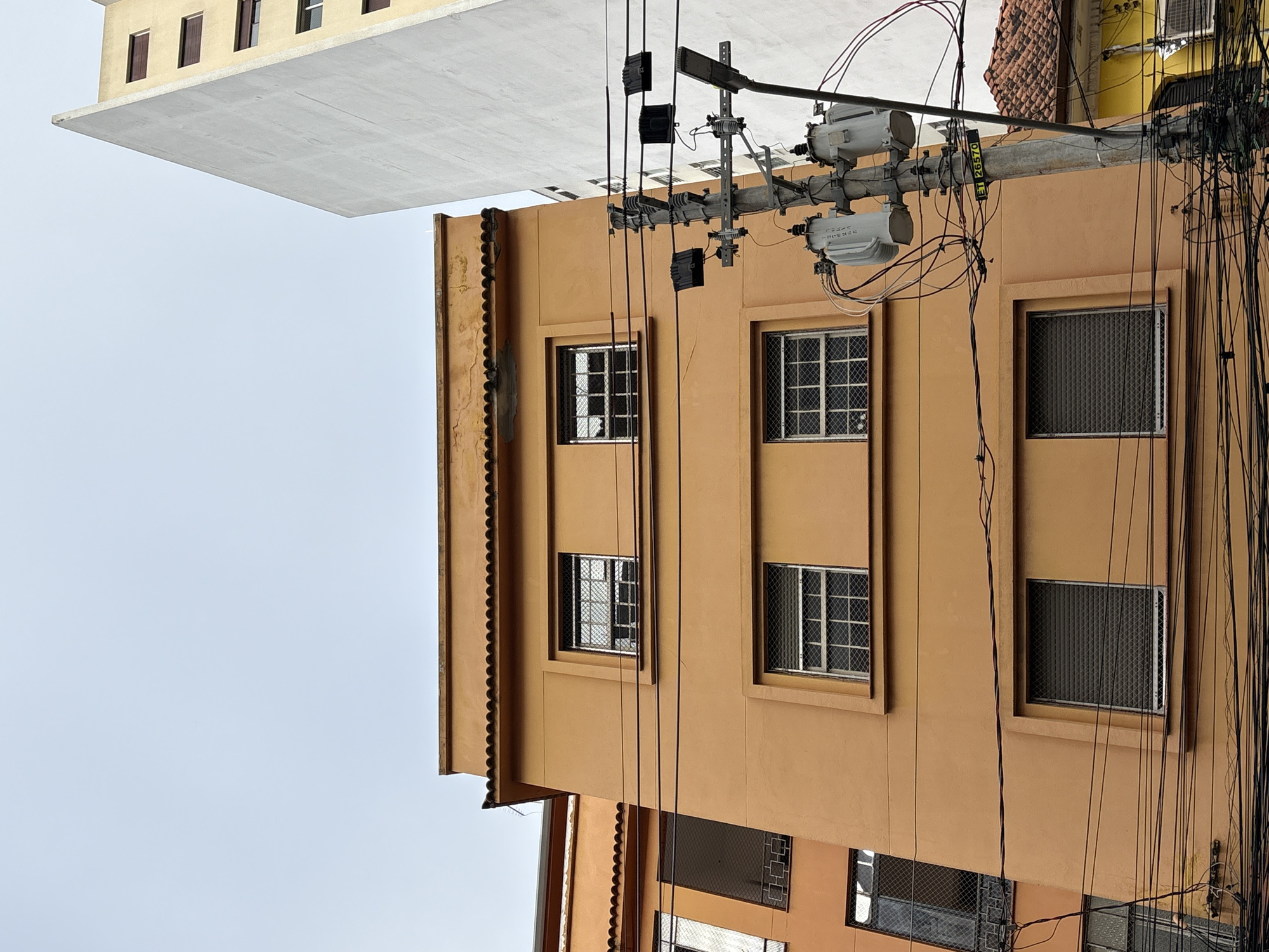 An eye-level, medium shot captures the exterior of buildings and a utility pole in São Paulo, Brazil. The scene is dominated by an orange building with multiple windows, some covered with metal grates. To the right, a tall, white building stands adjacent to the orange structure, partially obscuring it. A utility pole, adorned with transformers and wires, is positioned in the foreground, adding a sense of urban complexity to the scene. The sky above is a pale blue, suggesting an overcast day. The composition suggests a dense urban environment, typical of a bustling city.