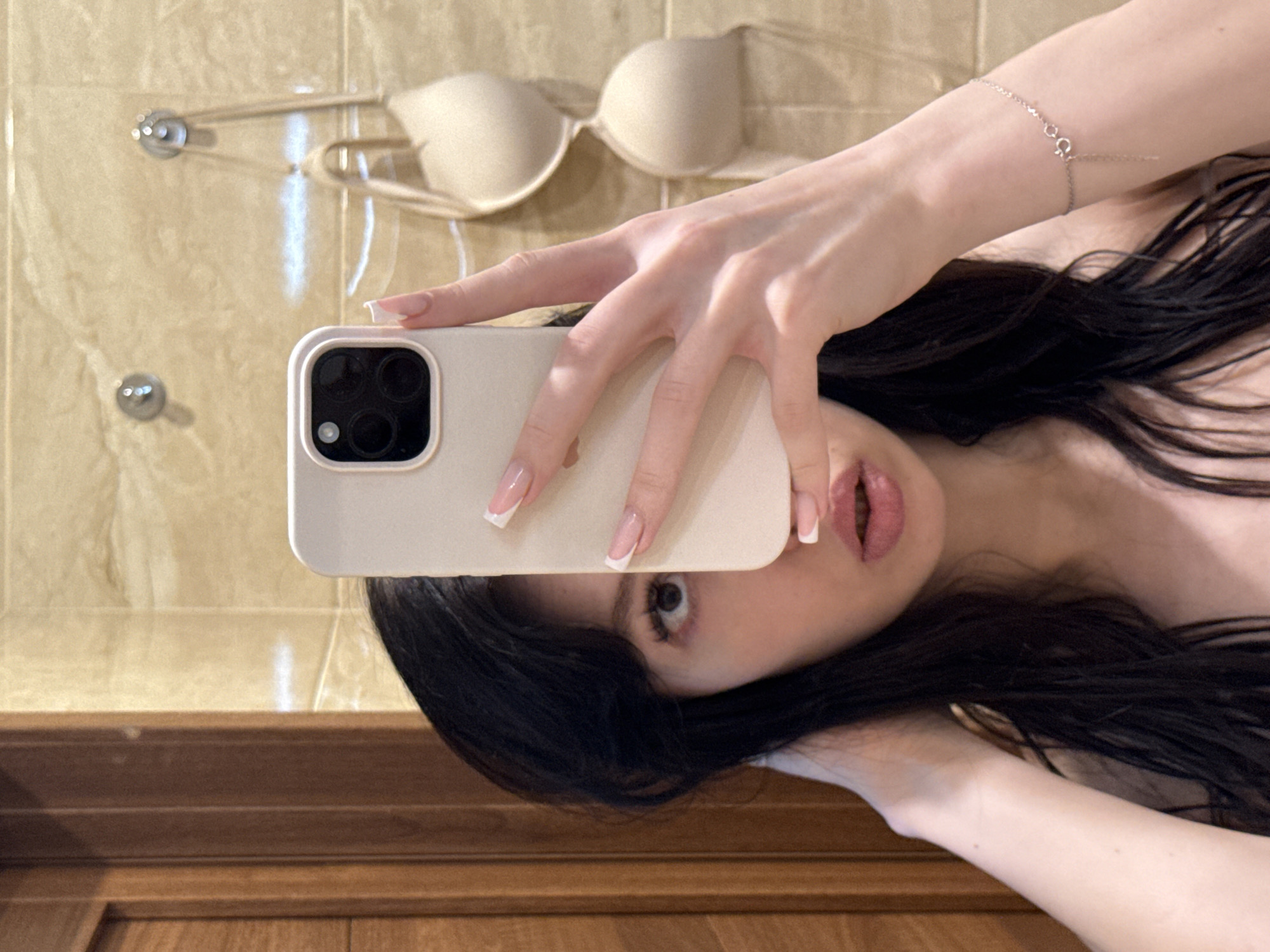 In a bathroom with beige tiled walls and a wooden door, a young woman with long, dark hair is taking a selfie. She is holding a white smartphone in her right hand, obscuring part of her face. She is wearing a light-colored bra, with the straps visible. The background shows two beige bras hanging on hooks. A delicate silver bracelet with a clasp is visible on her left wrist.