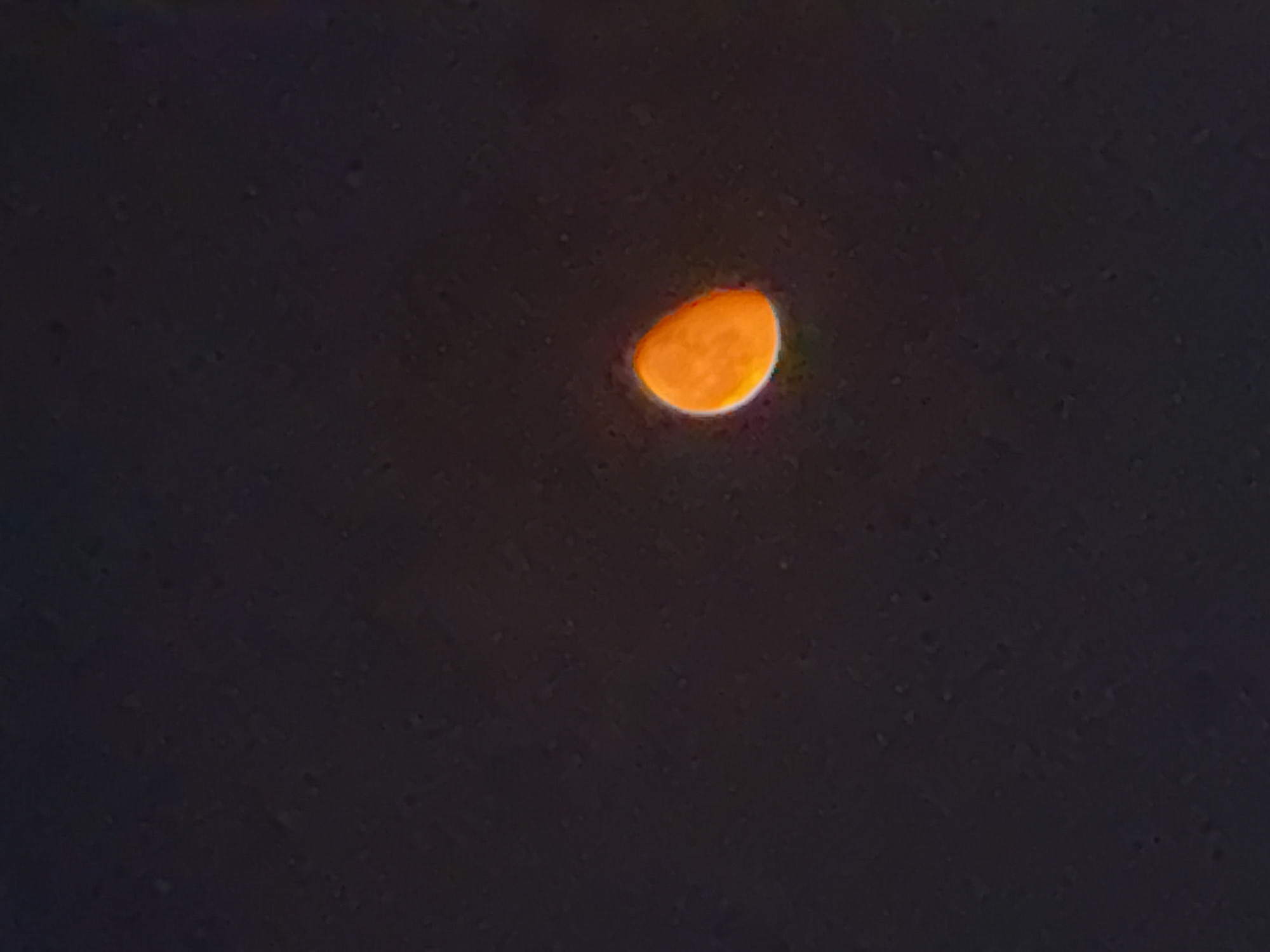 The image depicts a partial view of the moon against a dark sky. The moon appears as a crescent shape, illuminated with a vibrant orange hue, and edged with a soft, white glow. The background is predominantly dark and speckled, suggesting a night sky with some atmospheric haze or distant light sources. There are no visible landmarks, structures, or people that would indicate a specific location within Tbilisi, Georgia. The composition focuses solely on the celestial body and its illumination.
