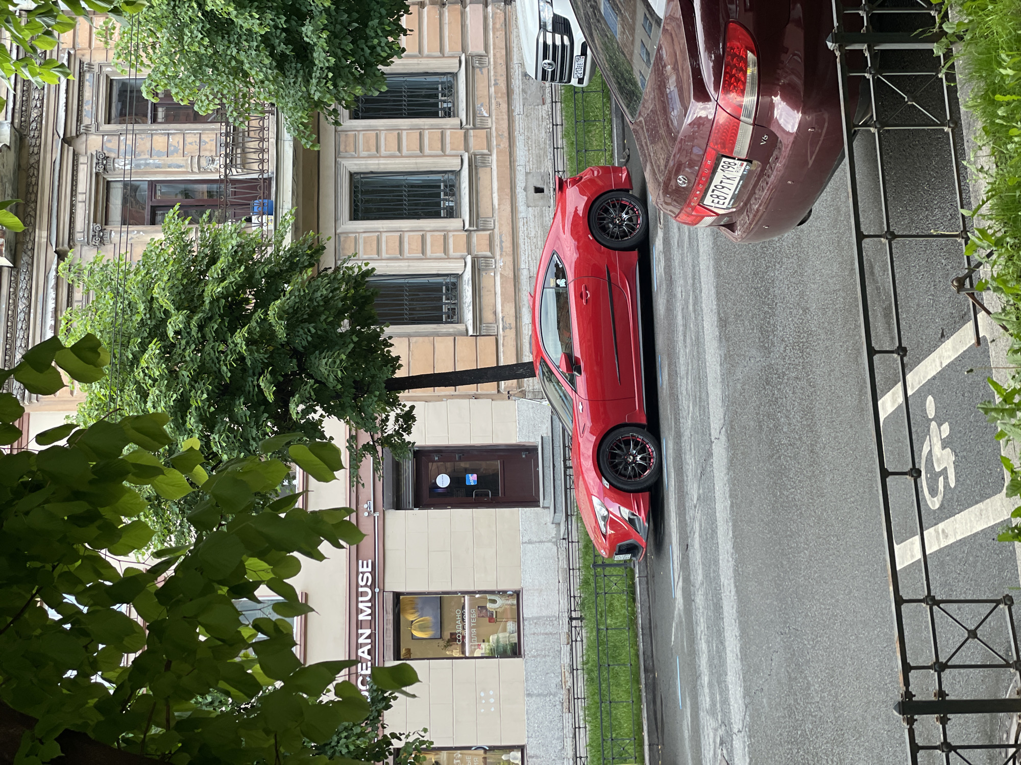 This media file captures an urban street scene in Saint Petersburg, Russia, during daytime under what appears to be overcast conditions. The image is composed of a historical building, two parked cars, and lush green foliage.
On the left side of the frame (when correctly oriented), a multi-story building with ornate classical architecture is visible. It features decorative cornices, pilasters, and windows with intricate grates. The ground floor houses a storefront, clearly displaying the English text "OCEAN MUSE" and partial Russian text "СОЗДАНО ДЛЯ ТЕБЯ" (Created for you) in its window.
The street, made of asphalt, occupies the right side of the image. Two cars are parked along the curb. The first is a vibrant red sports car, possibly a Subaru BRZ or Toyota GT86, notably featuring black aftermarket wheels with red accents. Behind it, a burgundy Hyundai Grandeur V6 sedan is parked. Both cars display Russian license plates with the "198 RUS" region code, confirming the Saint Petersburg location. Specifically, the red car's plate shows "O703EE 198 RUS" (partially visible on the white car in the crop) and the burgundy car's plate reads "E879TK 198 RUS".
The street also features a painted handicapped parking symbol and blue demarcation lines. A dark metal fence runs along the sidewalk, separating the road from a strip of grass. Abundant green trees and foliage frame the scene, with some leaves prominent in the immediate foreground and others partially obscuring parts of the building and the sky. No people or specific activities are visible, presenting a static street view.
