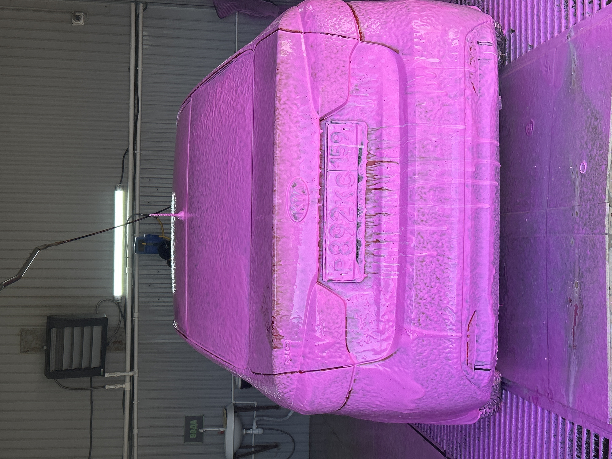 The image depicts the rear of a pink Kia Rio covered in pink foam, likely during a car wash. The license plate reads "B892KC 159", indicating the car is registered in the Perm Krai region of Russia. The backdrop is a corrugated metal wall and industrial lighting fixtures. A sign with Cyrillic text "ВОДА" (Water) is visible. The location is possibly a car wash facility in the city of Chaikovsky, Russia, given the license plate region. The car is positioned on a metal grate floor.
