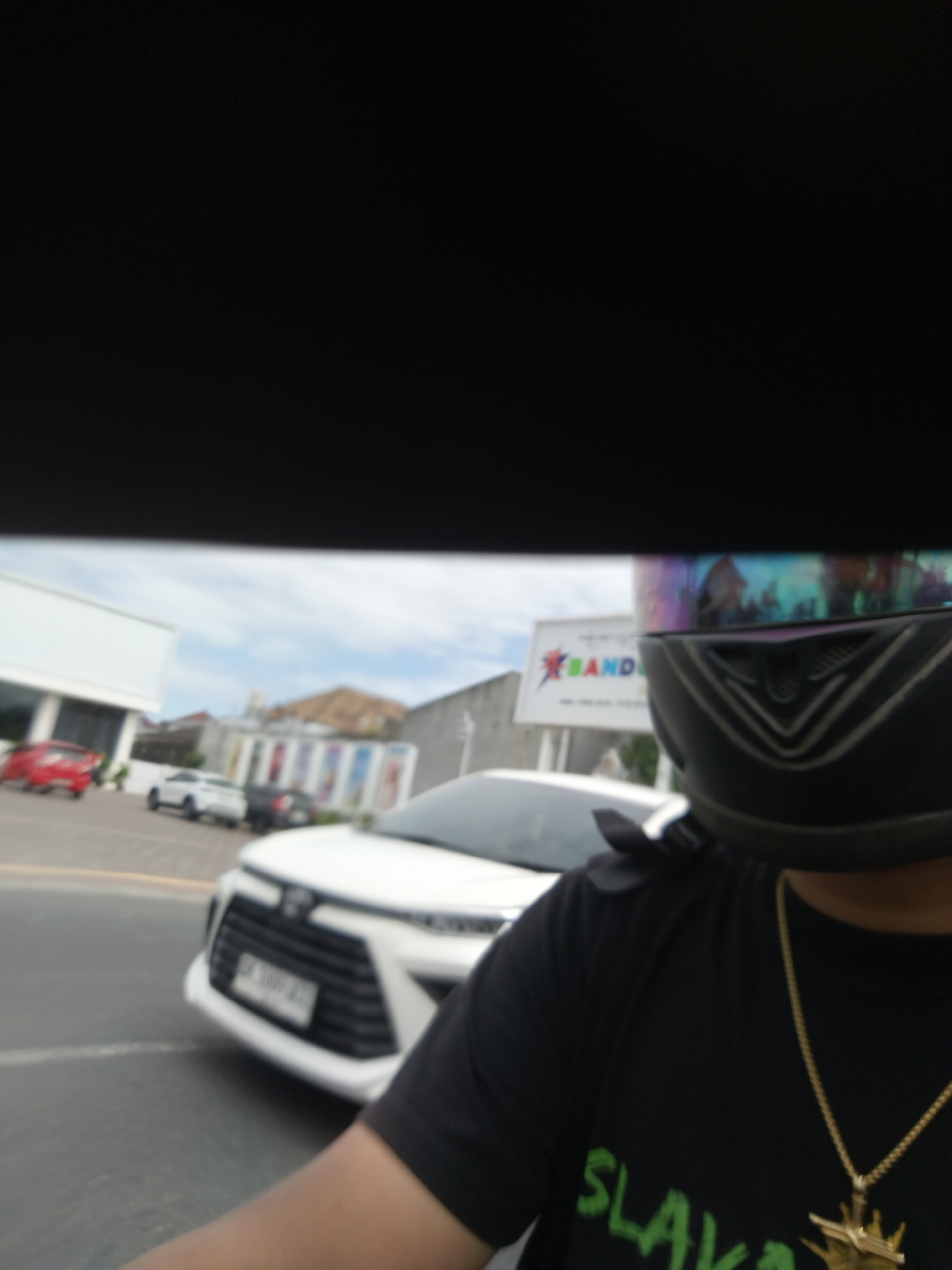 The image shows a person wearing a black t-shirt and a black helmet with a iridescent visor, taken from a first-person perspective. The person is wearing a gold chain with a pendant. The background suggests a street scene in Denpasar, Indonesia. A white car with its front visible is in the foreground, partially obscuring the person. In the background, there are other cars, a building with a white facade, and a billboard with the word "BAND" visible on it, along with what appears to be colorful vertical stripes on another building. The sky is cloudy. The overall impression is a candid shot taken while riding a motorcycle or similar vehicle.