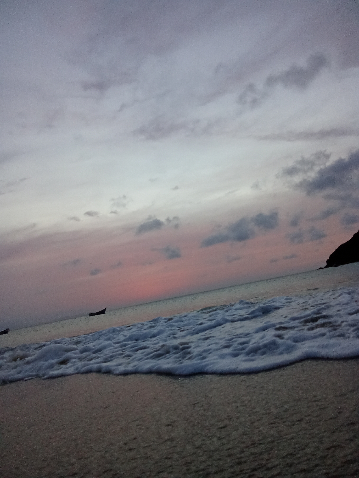 The image depicts a beach scene in Juan Griego, Venezuela, at dusk or dawn. The sky is a mix of grey and pink clouds. The sea is visible with foamy waves reaching the shore. Two small boats are silhouetted on the horizon. A dark headland is seen on the right. The beach sand occupies the foreground. There are no people visible in the image.
