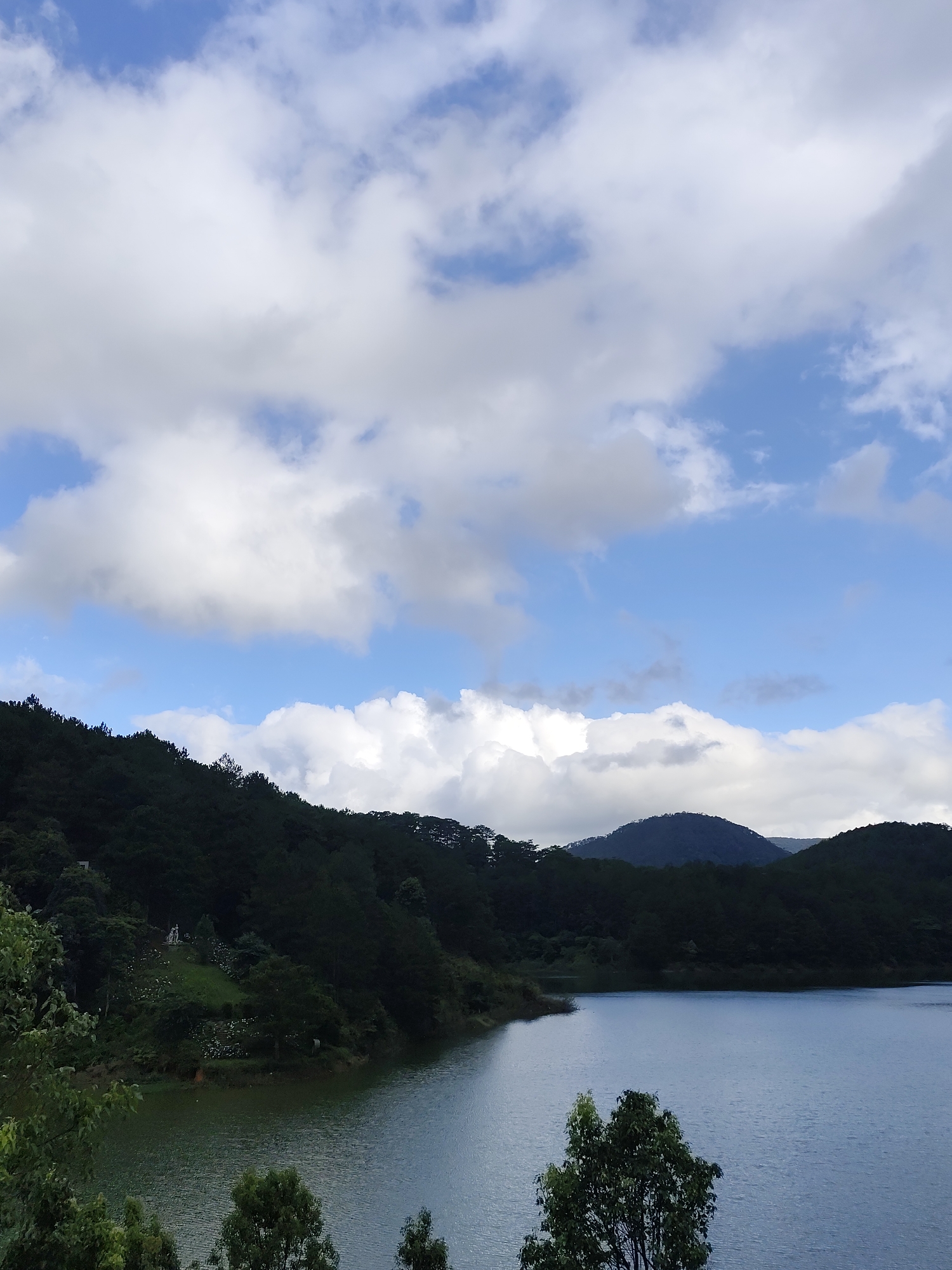 The image depicts a lake surrounded by lush, green hills, likely within or near the city of Da Lat, Vietnam. The sky is predominantly blue, dotted with large, white cumulus clouds. The hillsides are densely covered in trees, creating a dark green backdrop to the lake. The lake's surface is generally smooth. The foreground includes trees. In the distance, there are hazy hills that extend toward the horizon. A small clearing with a light structure is visible on the near hill.