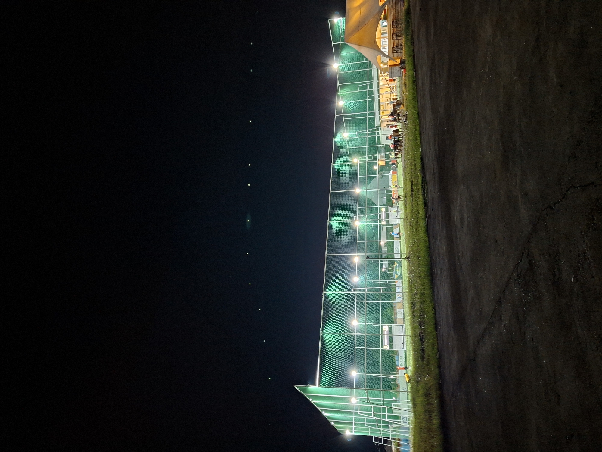 The image shows a night scene in Lhokseumawe, Indonesia. A fenced and illuminated sports facility, possibly a badminton or futsal court, borders a paved area. The court is lit by evenly spaced lamps. Beyond the court, a building with a partially visible structure, possibly a cafe or restaurant, features overhead lighting and figures seated at tables. The foreground pavement stretches into the right side of the frame. The left portion of the image is dominated by a dark, unlit sky. There is no visible activity on the court itself.