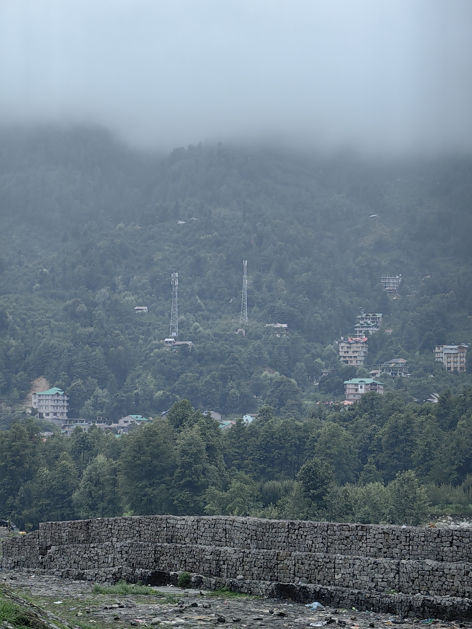 The image depicts a view of Bahang, India, characterized by a hillside covered in dense green foliage, with scattered buildings and communication towers visible. In the foreground, a layered stone wall runs horizontally across the frame, with a ground surface littered with debris, including plastic waste and stones. The sky is overcast and hazy, obscuring the upper portions of the mountains. The buildings are multi-story structures, some appearing to be hotels or residences, interspersed among the trees on the slopes. Communication towers stand prominently on the hill. The overall atmosphere is muted and slightly damp, suggesting a cloudy or misty day. No people are visible in the frame.