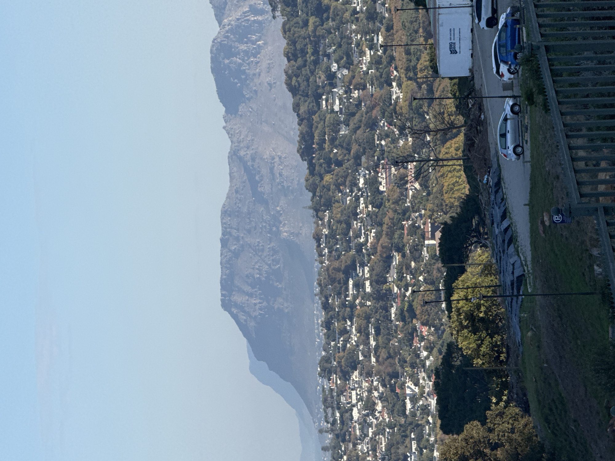 The image shows a view of Cape Town, South Africa, with Table Mountain visible in the background on the left side. A dense residential area with houses and trees stretches out in the mid-ground. A road runs along the right side of the image, featuring parked cars, a moving company truck, lampposts, and a retaining wall bordering the road. The sky is clear and light blue.