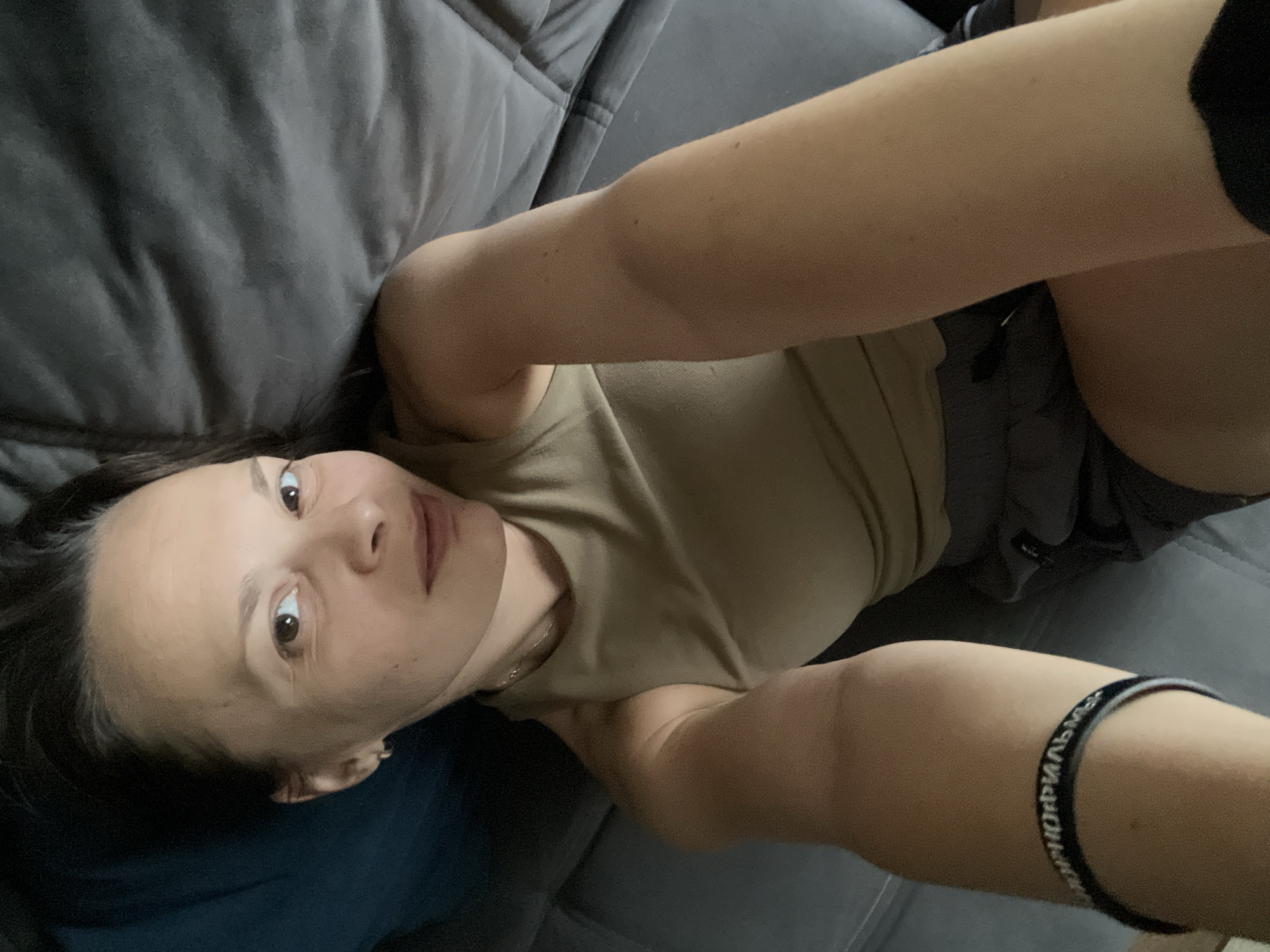 A woman with dark hair and brown eyes lies on a gray sofa, looking at the camera. She is wearing a sleeveless tan top and dark shorts. A black bracelet with white text is visible on her left wrist. The background consists of the sofa and a dark blue pillow. The scene appears to be an indoor setting, possibly a living room, in Tolyatti, Russia.