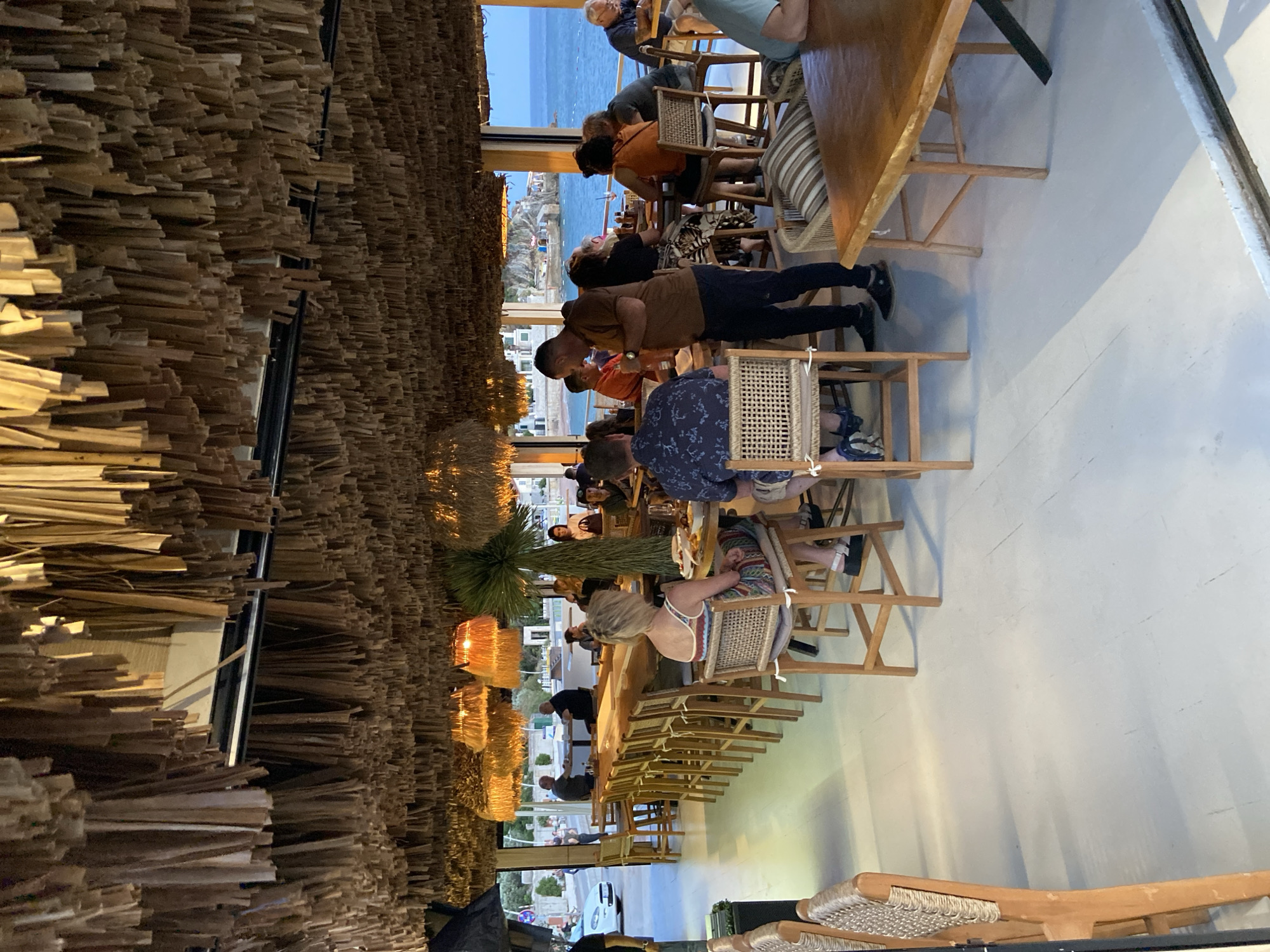 The image depicts a restaurant interior in Petrovac centar, Montenegro. The ceiling is covered in a dense arrangement of wooden sticks, creating a textured effect. A palm tree stands near the center of the scene, and hanging lamps provide warm light. People are seated at long wooden tables, seemingly dining or conversing. The restaurant opens to a view of the sea and coastline through a large window. Chairs are made of wood with woven seats.