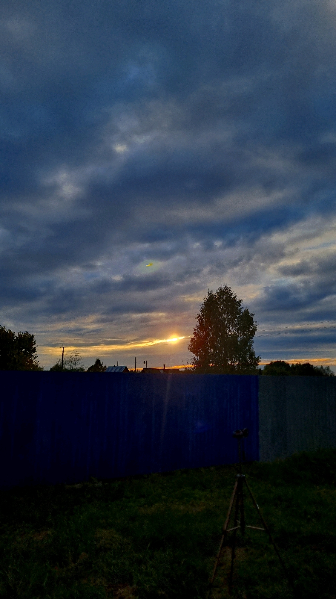 This image captures a sunset over the city of Vysokoe, Russia. The sky is dominated by dark, textured clouds, with a sliver of bright orange sunlight breaking through near the horizon. A lone tree stands silhouetted against the vibrant sunset, to the right of the frame. In the foreground, a dark blue fence stretches across the lower half of the image. Beyond the fence, hints of houses and utility poles are visible. To the right of the fence, a tripod with an optical device is set up on a patch of green grass, suggesting an observation or photography activity. The overall mood is serene and atmospheric, typical of a twilight scene.