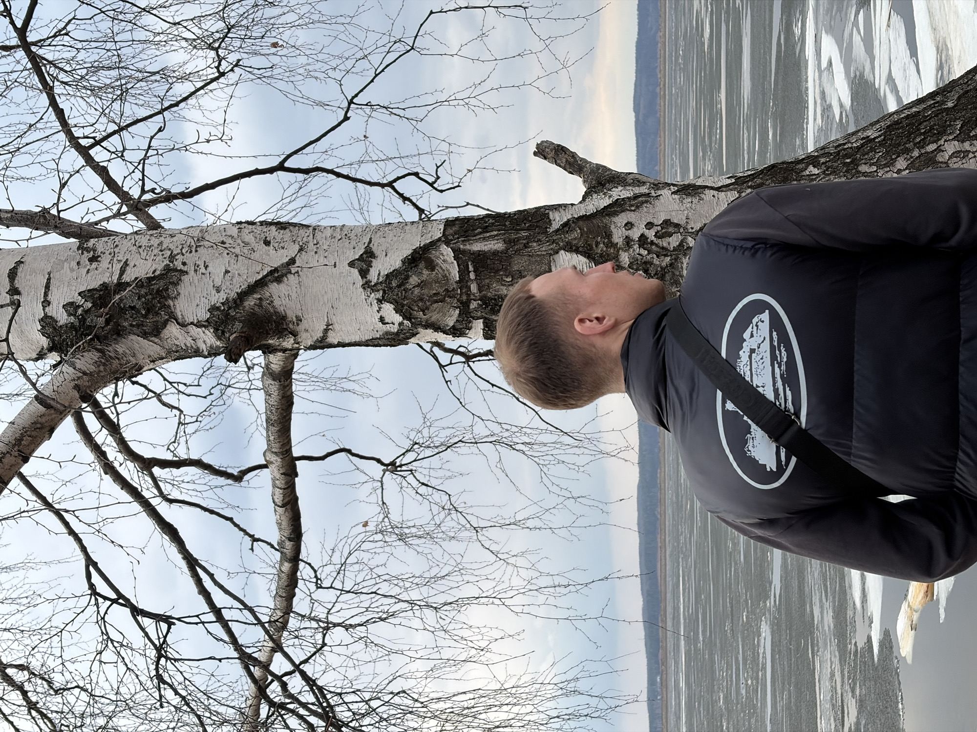 The image shows a man standing next to a birch tree. The man is wearing a black jacket with a white logo on the back and a shoulder strap. He is looking up at the tree. The birch tree has a white trunk with black markings, and its branches are bare. In the background, there is a view of a frozen body of water and a distant shoreline. The image was taken in Chaikovsky, Russia.