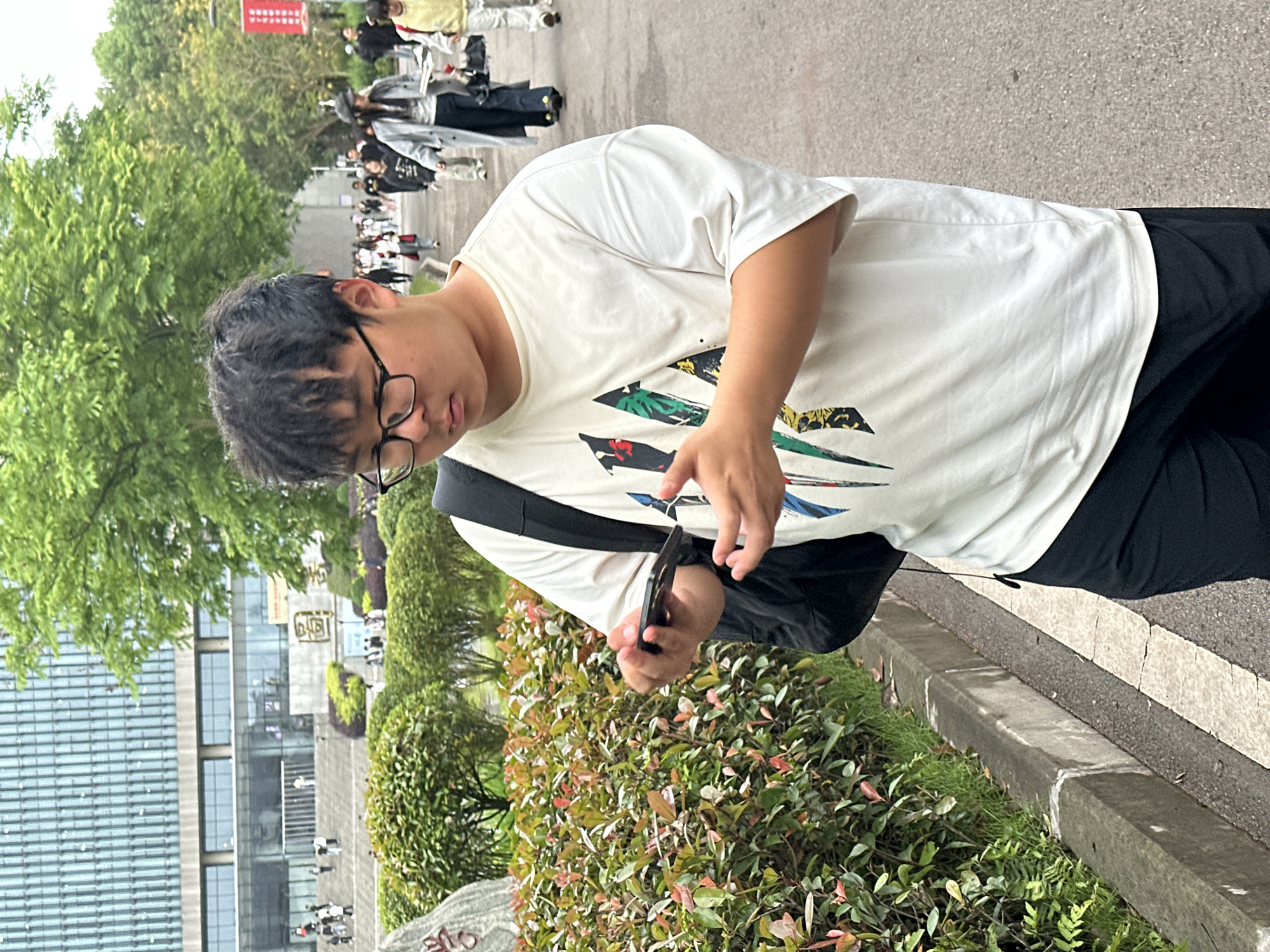 A person wearing a white t-shirt with a graphic design and glasses is standing outside, looking at a black smartphone in their hands. A black bag is slung over their shoulder. In the background are trees, a building with glass windows, and a crowd of people. The location is likely in Jiangjin, China. The person is standing next to a curb and bushes.
