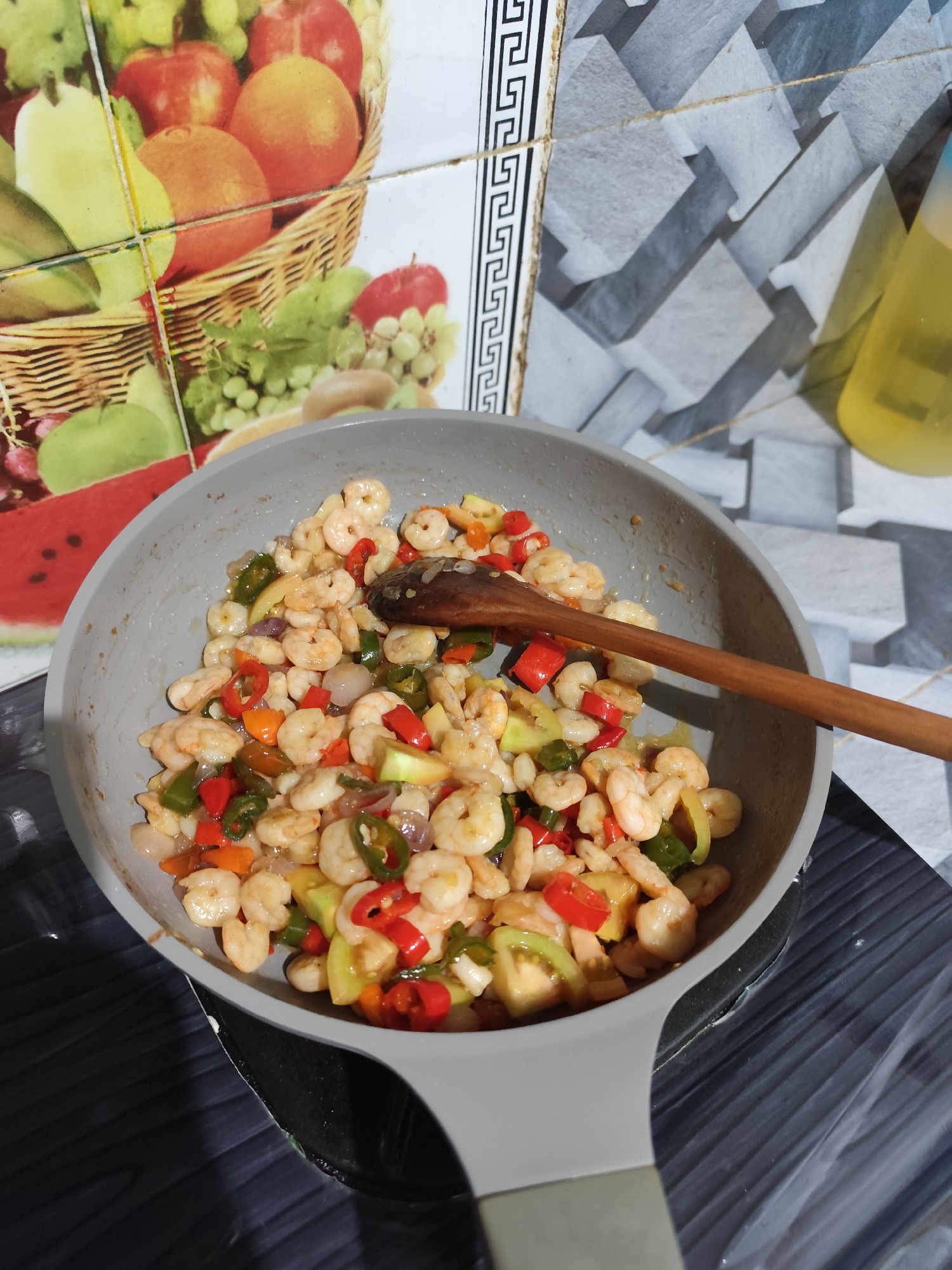 An overhead shot shows a close-up of a light gray frying pan filled with cooked shrimp and diced vegetables. The shrimp are small and pinkish-white, and they are mixed with chunks of red bell pepper, green chili peppers, and some lighter green vegetables. A wooden spoon rests on top of the contents of the pan, its handle extending towards the right side of the frame. The pan is placed on a black stovetop, which has a textured surface. The background features a tiled wall with a fruit basket design on the left and a geometric pattern on the right. A yellow bottle is visible in the upper right corner. The scene appears to be in a kitchen.