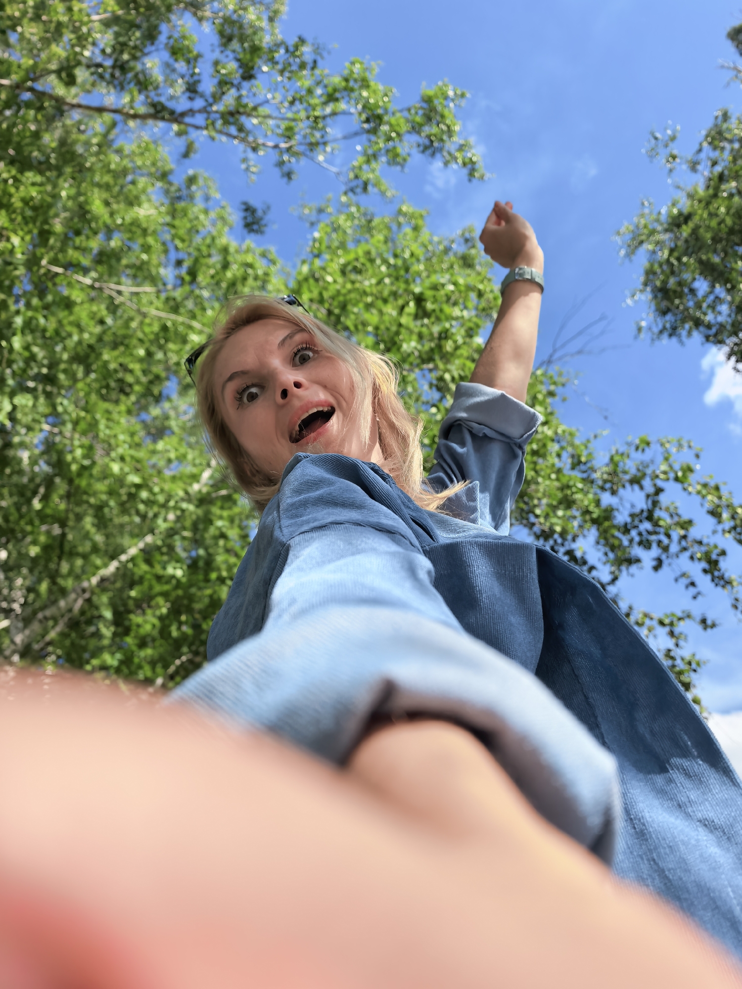 The image depicts a low-angle perspective of a blonde-haired female looking down directly at the camera in an outdoor setting within the city of Sterlitamak, Russia. Her eyes are wide, and her mouth is open in an expression suggesting surprise or playfulness. Sunglasses are perched on top of her head. She wears a blue, textured long-sleeved shirt or jacket, possibly corduroy, with the sleeves rolled up. Her right arm is raised and bent at the elbow, displaying a light green wristband or watch. The background features a clear blue sky with sparse white clouds and numerous vibrant green leaves from deciduous trees, indicating a sunny day. A heavily blurred, reddish-pink mass, consistent with fingers or a hand, occupies the immediate foreground at the bottom-left, suggesting the camera was held by or very close to the subject's hand.