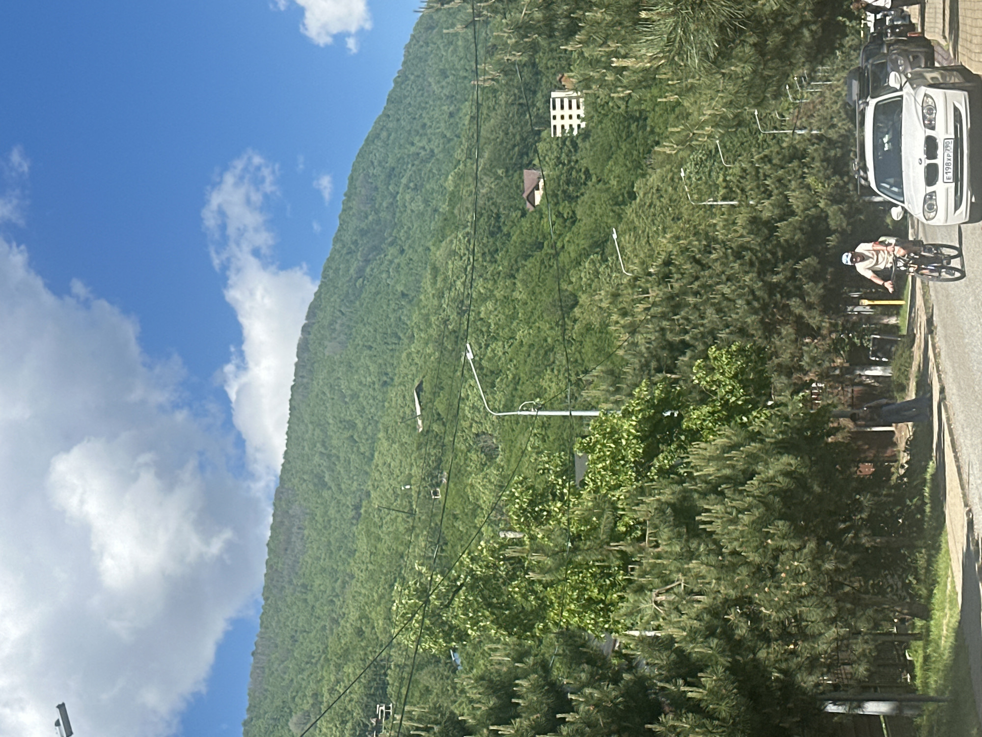 The image captures a vibrant daytime scene in a mountainous area, likely Krasnaya Polyana, Russia, under a bright blue sky with scattered white clouds. A steep, densely forested green mountain or hillside dominates the left and central parts of the frame, showcasing lush vegetation.
On the right side of the image, a paved road is visible, along which a person on a bicycle is riding. The cyclist wears a light-colored shirt, shorts, and a helmet, and is pedaling towards the background. Parked cars line the road, including a white BMW with the visible Russian license plate "E198XP 190." Other darker vehicles are parked further behind the BMW. Streetlights and utility poles with overhead wires run parallel to the road and extend into the mid-ground.
Several small buildings are nestled within the green expanse of the hillside. One notable structure is a white building with a distinctive grid-patterned facade, and another with a dark, possibly brown, roof is partially visible below it. The foreground features tall, coniferous trees, providing depth to the scene. The weather appears sunny and clear, suggesting a pleasant day. The activity primarily involves the cyclist and parked vehicles, set against a backdrop of natural beauty and some human development.