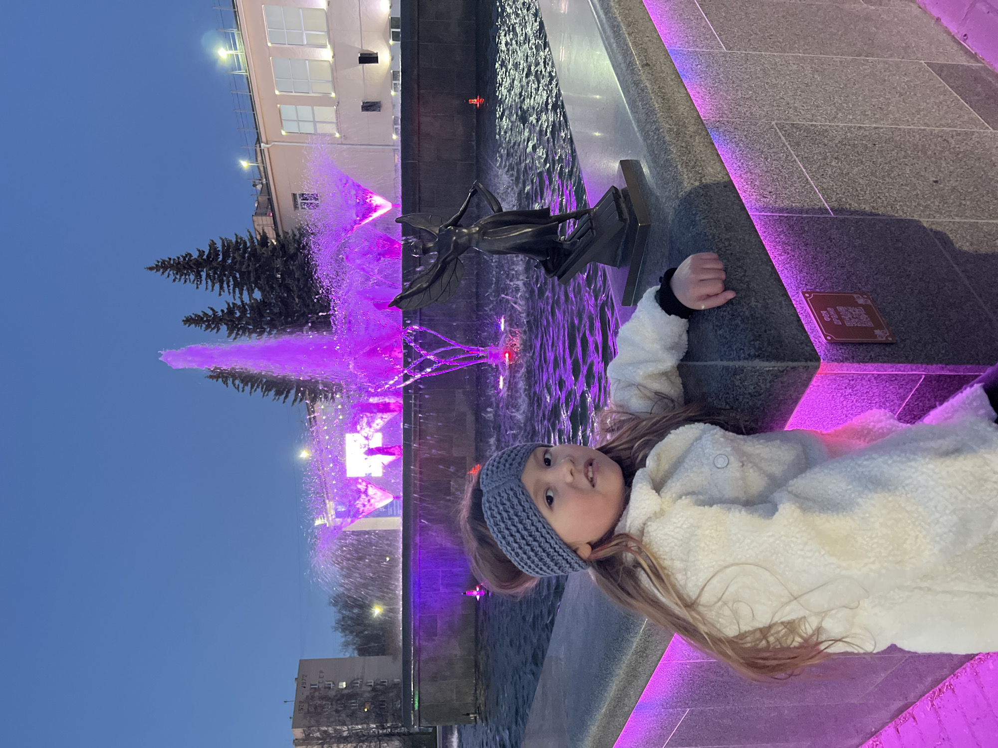 A young girl lies on the edge of a fountain in Chaikovsky, Russia. She wears a gray knit headband and a white fleece jacket. The fountain has a statue of a winged figure and features purple lighting. Water is spraying from the fountain. Buildings are visible in the background.