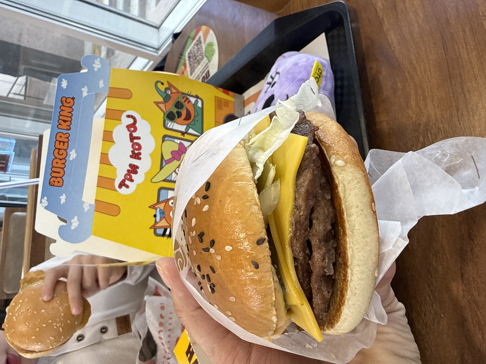 This indoor shot captures a close-up of fast food items on a wooden table, likely in a Burger King restaurant in Tobolsk, Russia. The primary focus is a large cheeseburger held in a person's hand, partially wrapped in white paper. The burger features a sesame seed bun, a thick meat patty, two slices of melted yellow cheese, and shredded lettuce. The bun has both white and black sesame seeds.
In the background, on the left, a smaller burger is visible, held by what appears to be a child's hand, suggesting a meal enjoyed by more than one person. Behind these burgers is a colorful cardboard box or container, likely a kids' meal box, featuring cartoon characters. Visible text on this box includes "BURGER KING" in orange letters on a blue section, and "ТРИ КОТОЯ" (which translates to "Three Cats") in white speech bubbles on a yellow background, confirming the Russian context.
The items rest on a black plastic tray. A purple plush toy, possibly a character from the "Three Cats" show, with a yellow tag, is also visible on the tray to the right of the cardboard box. The background shows glimpses of a window or glass partition, reflecting light. The overall scene suggests a casual dining experience at a fast-food establishment.