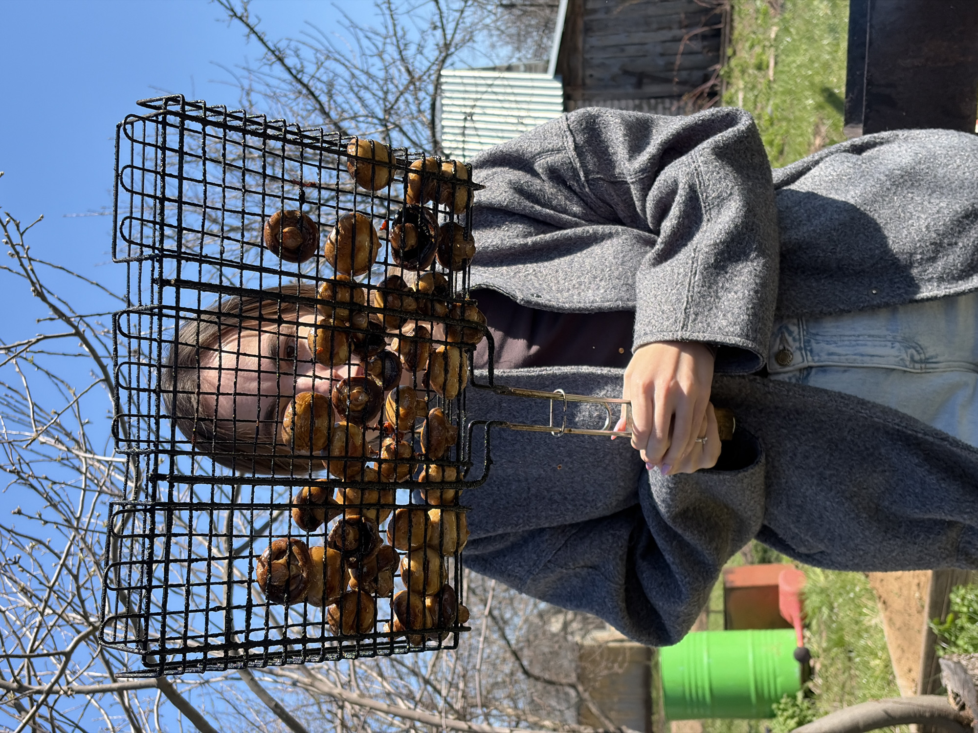 A person in a gray coat and blue jeans stands outdoors, holding a black wire grill basket filled with cooked mushrooms. The background includes a clear blue sky, bare tree branches, a wooden shed, and green grass. The location is suggested to be Neftekamsk, Russia, but this cannot be definitively confirmed from the image.