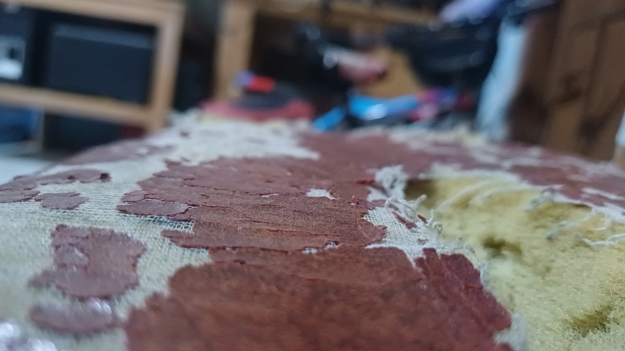 The image shows a close-up of a worn and torn upholstered surface, likely a chair or sofa, with a beige fabric base and reddish-brown peeling material. The texture is rough and fragmented, revealing yellow foam padding underneath in some areas. In the blurred background, a wooden shelf unit is visible, with a dark electronic device on it. Further back, other objects are indistinct, suggesting an indoor setting. The scene appears to be within a residential or storage space, with no immediate indicators pointing to a specific location in Wonokerso, Indonesia, beyond it being a domestic environment.