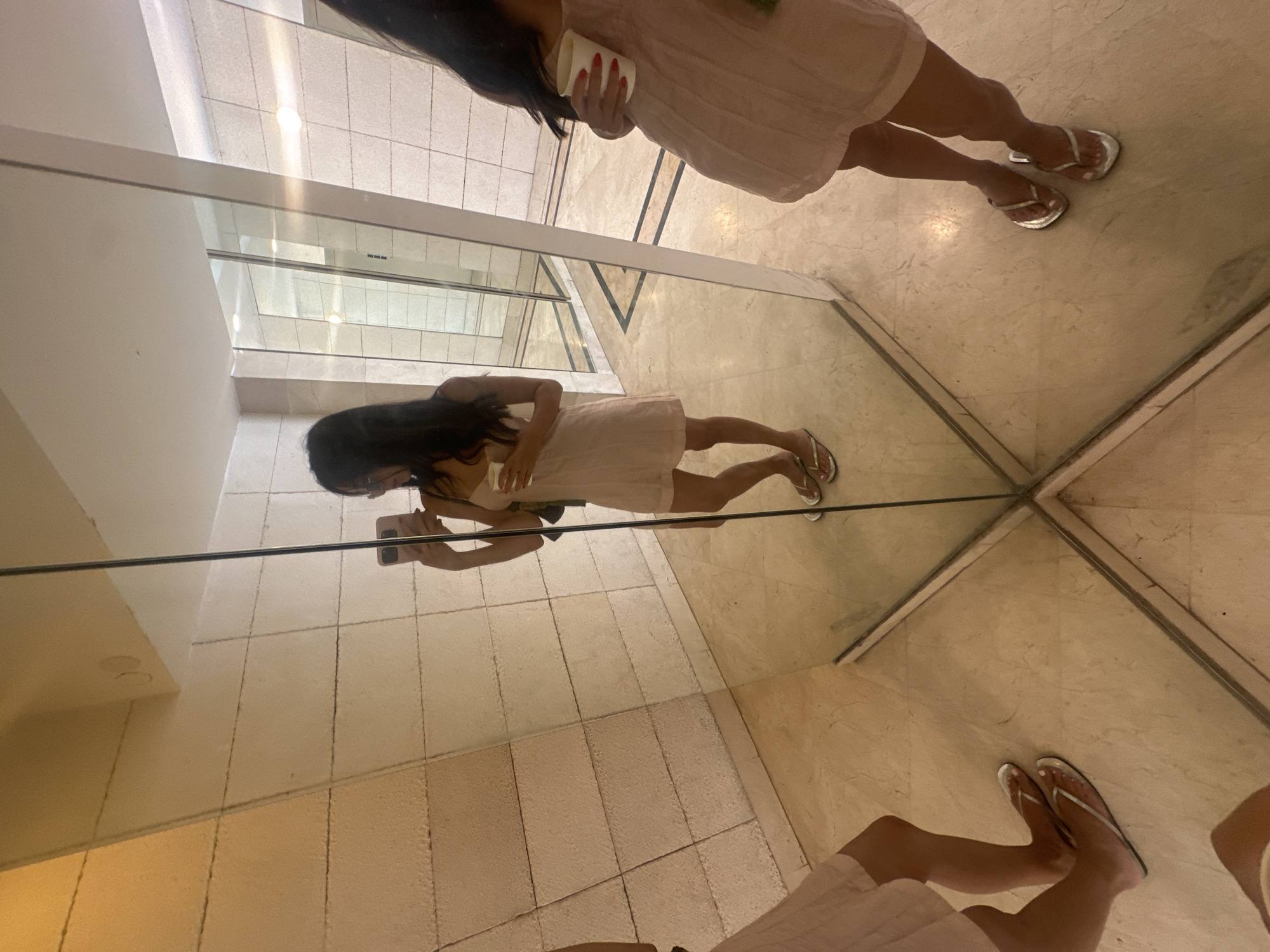 A woman with long dark hair takes a selfie in a mirrored room. She is wearing a light pink slip dress and silver flip-flops. She is holding a white paper cup in her right hand and a smartphone in her left hand. The room has tiled walls and a marble floor. In the reflections, it appears to be a hotel lobby or a similar public space. The background includes doorways and architectural details that are characteristic of modern buildings. The overall lighting is bright and ambient.