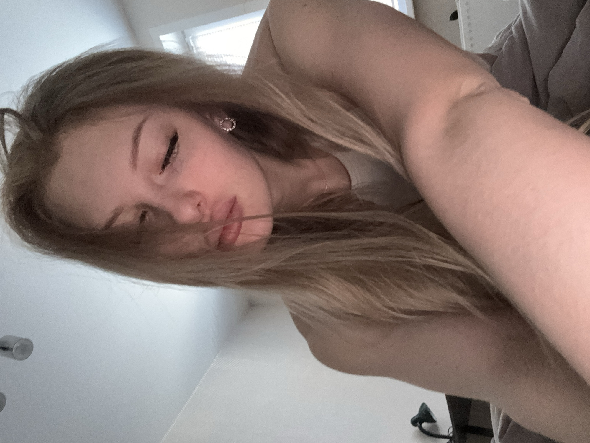 A fair-skinned woman with long blonde hair is lying down. She is partially facing the camera with her eyes closed and mouth slightly pursed. She is wearing a small, circular earring in her left ear. A thin necklace is visible around her neck. Her right arm is in the foreground, partially obscuring the view. The background includes a white wall and a window with blinds. A gray-colored object (possibly bedding) is also visible. The location is not discernible from the image.